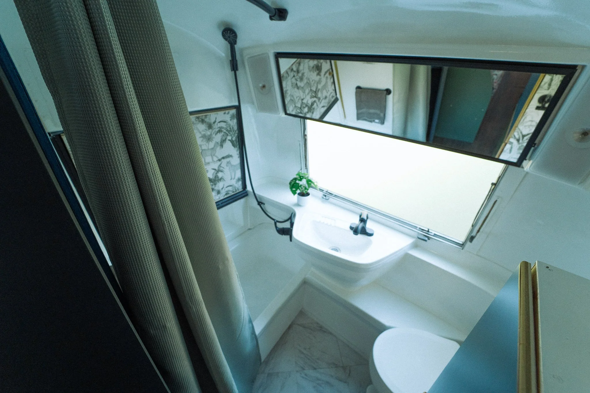 A small bathroom with an open frosted window, a white sink with a black faucet, a small green plant, a mirror above the sink, and cream-colored curtains.