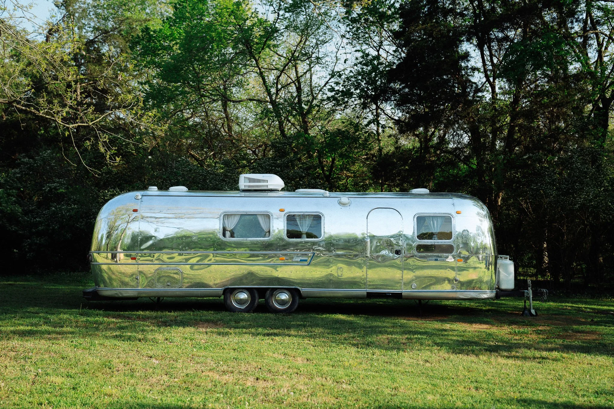 A shiny, metallic, vintage Airstream trailer parked on a grassy area with trees in the background.
