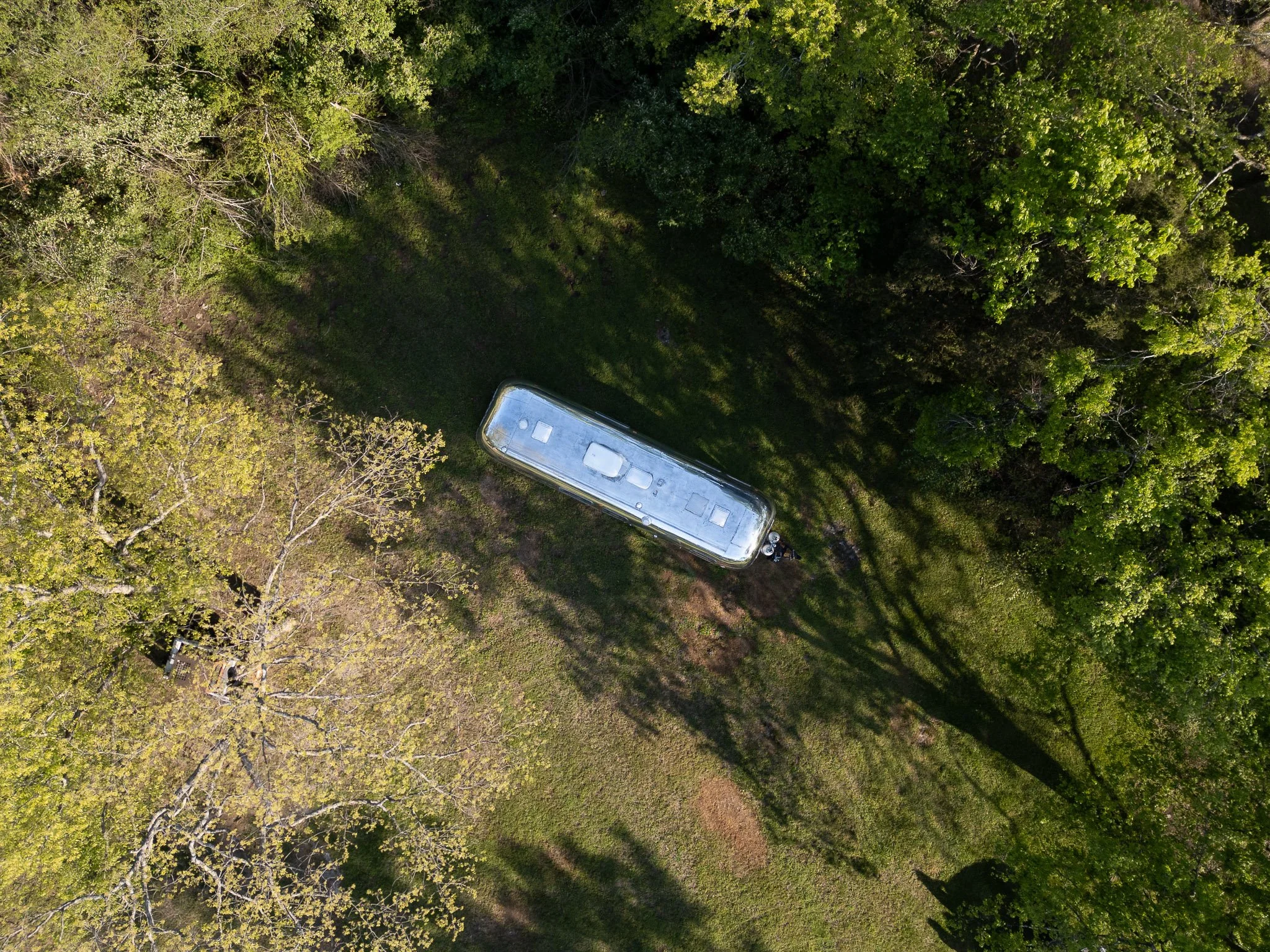 An aerial view of a silver camper trailer parked in a grassy clearing surrounded by trees.