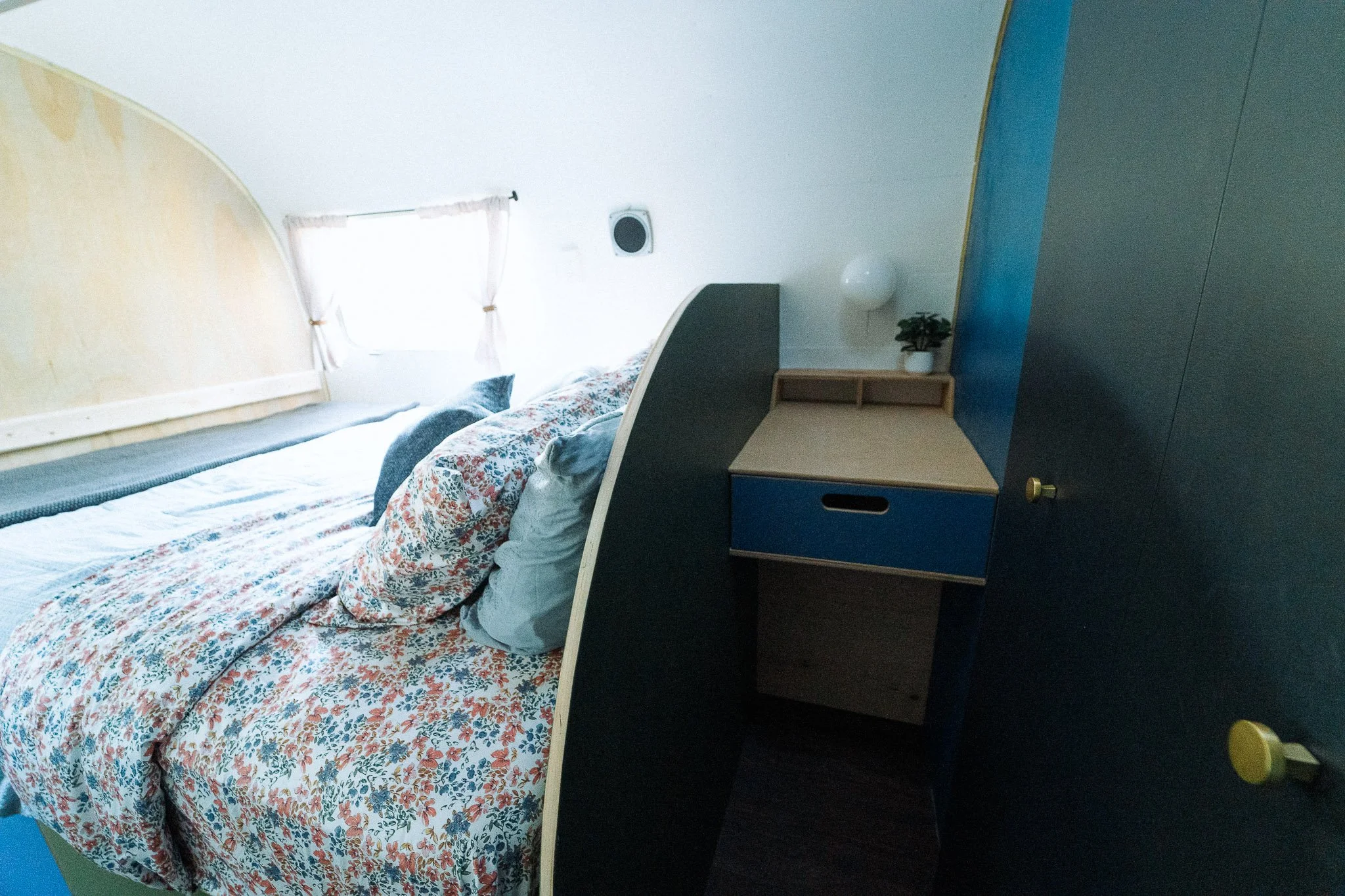 A cozy bedroom with a bed covered in a floral duvet, paired with various pillows. There is a window with white curtains, a blue and wooden nightstand with a small potted plant, a white wall-mounted lamp, and a dark wardrobe with yellow knobs.