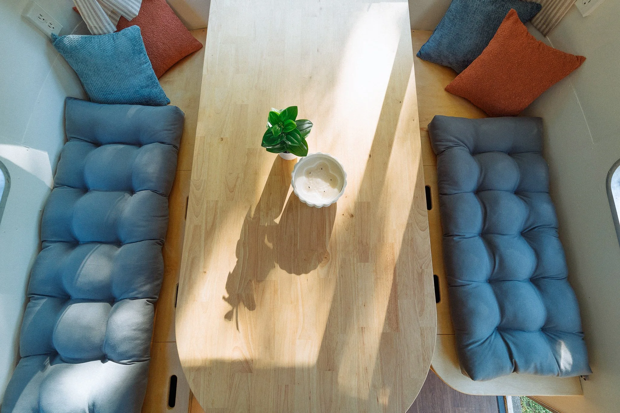 A top-down view of a dining table with a potted plant and a candle, surrounded by two blue cushioned benches with colorful pillows in a cozy kitchen or dining area.