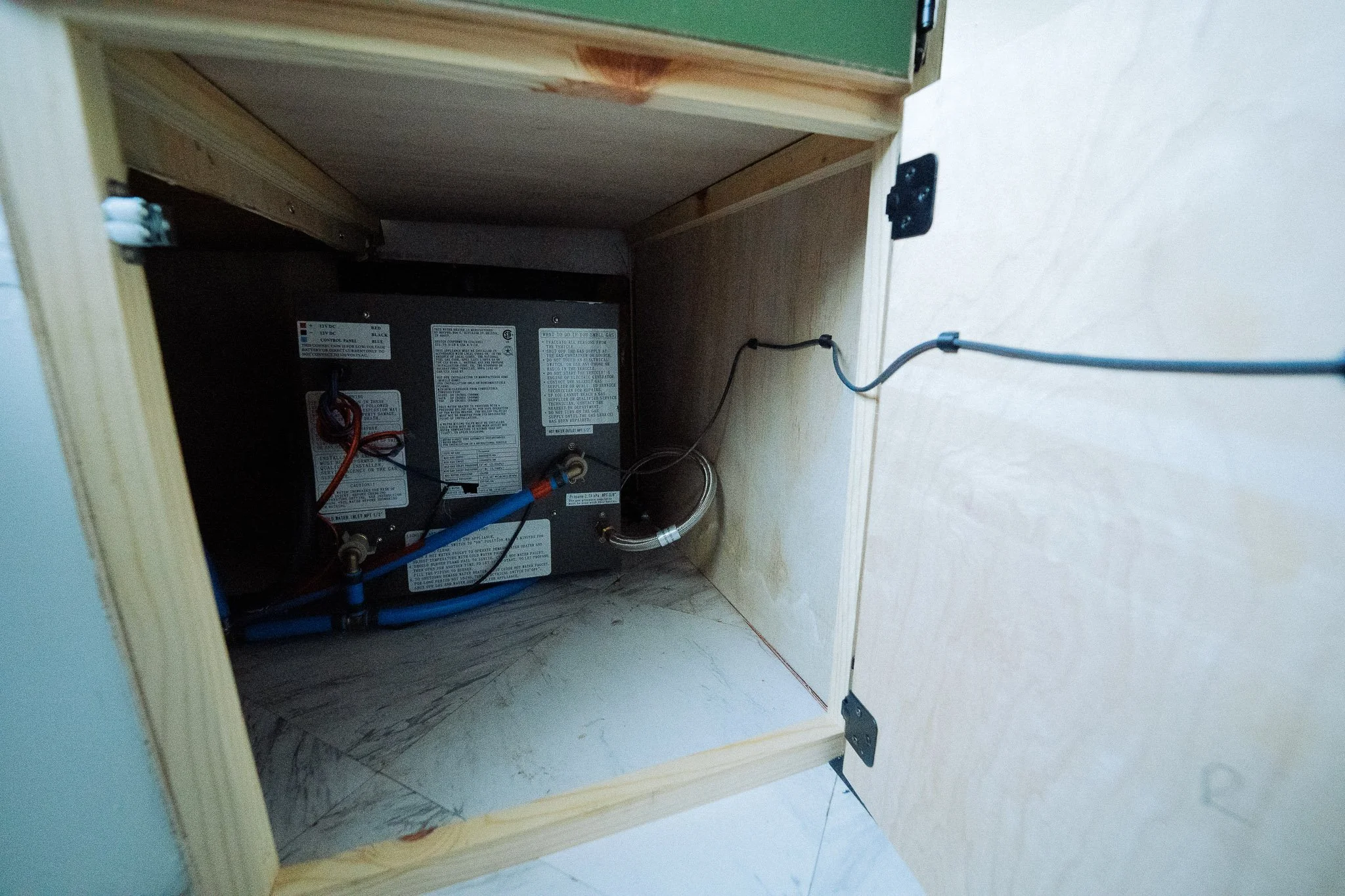 The inside of a cabinet with an electric unit, wiring, and pipes visible, set against a wall and tiled floor.