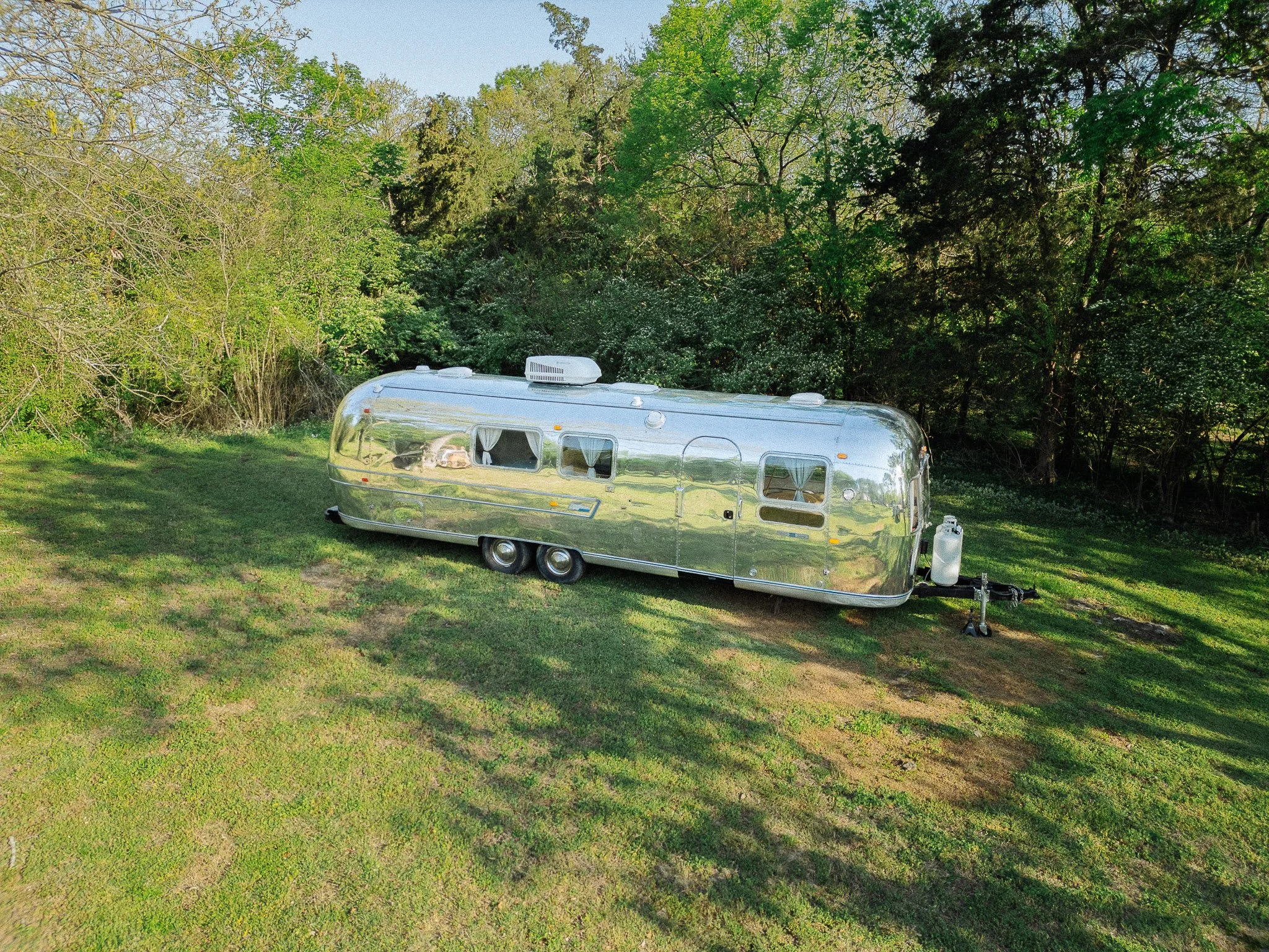 A shiny, reflective silver camper trailer parked on a grassy area with trees in the background.