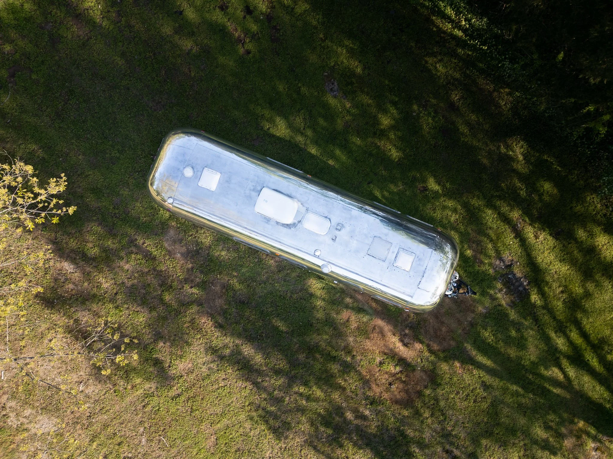 Aerial view of a shiny silver camper trailer on a grassy area surrounded by trees, with shadows cast on the ground.