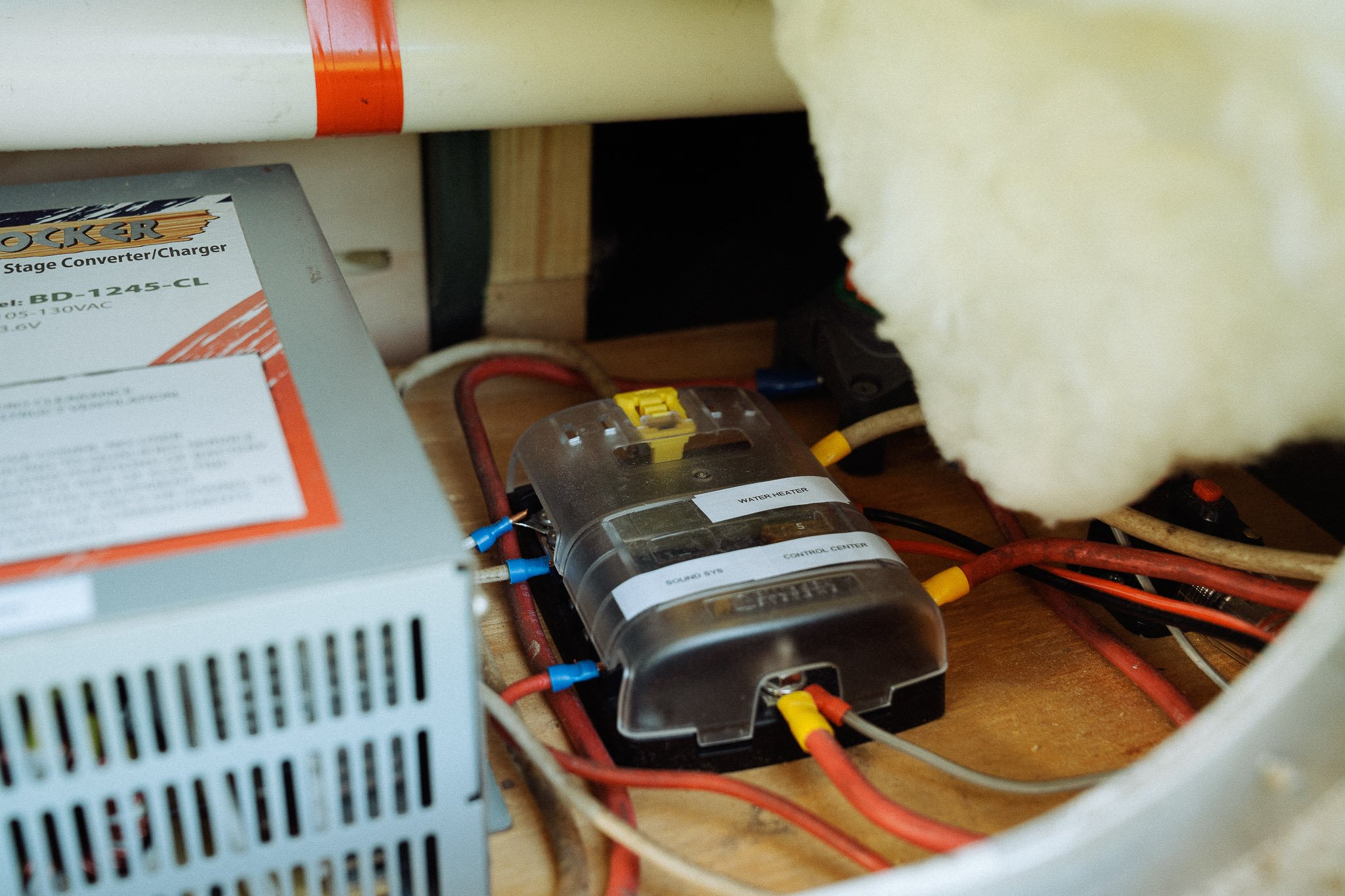 Close-up of electrical components in a confined space, including a metal power supply, a transparent water heater, and various wires, set against a wooden surface with insulation.