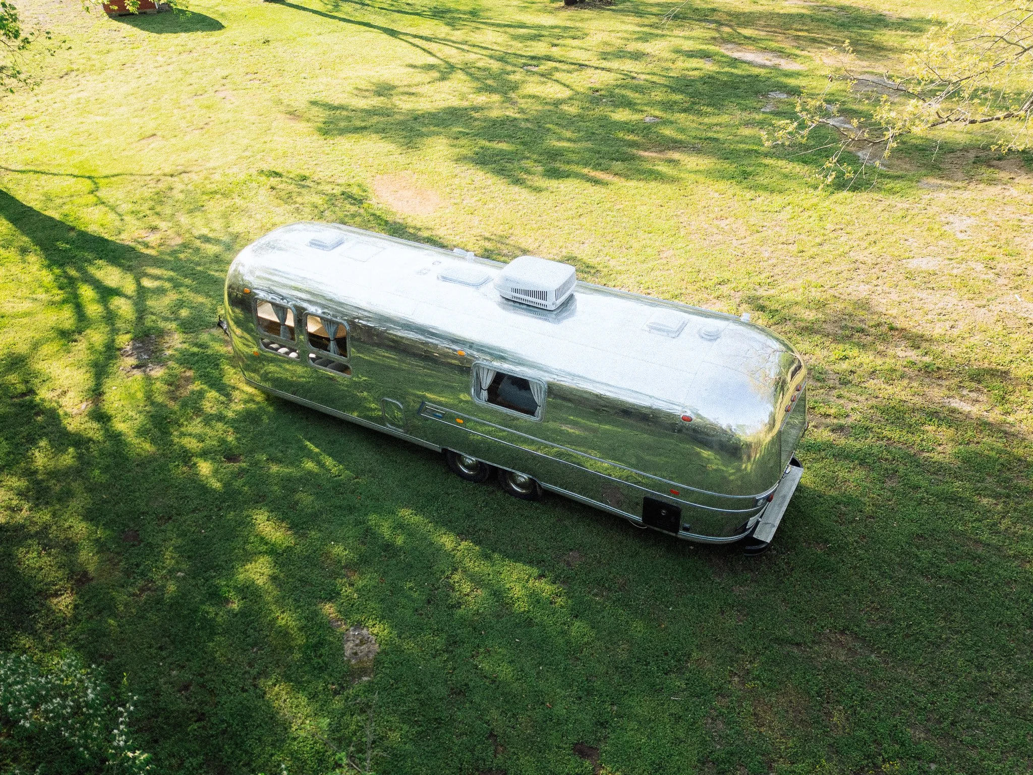 A shiny, silver Airstream trailer parked on a grassy field with trees, casting shadows on the ground.