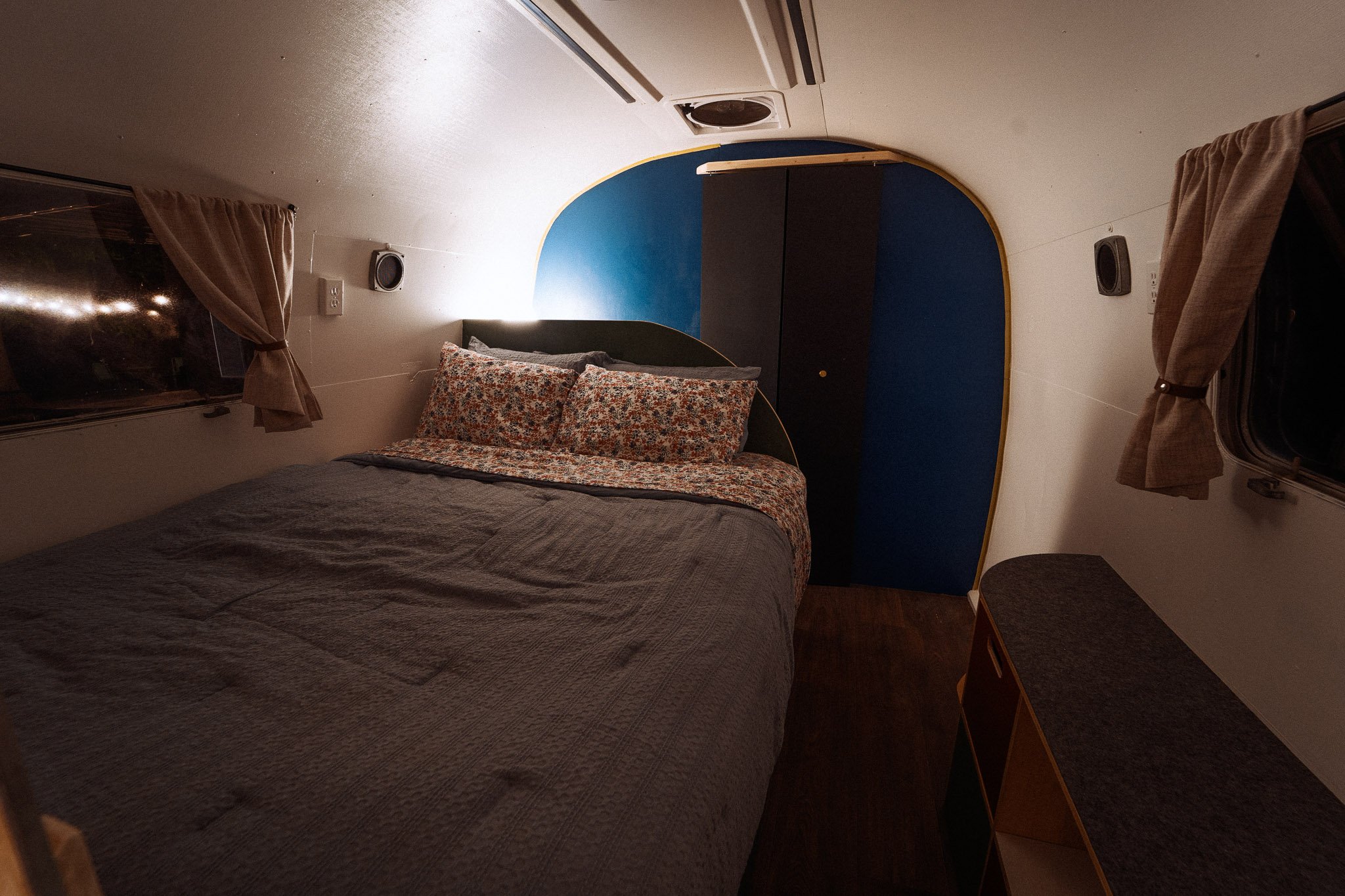 Interior of a small bedroom or sleeping area with a bed, floral-patterned pillows and bedding, small windows with curtains, a built-in wardrobe, and a dark wood dresser.