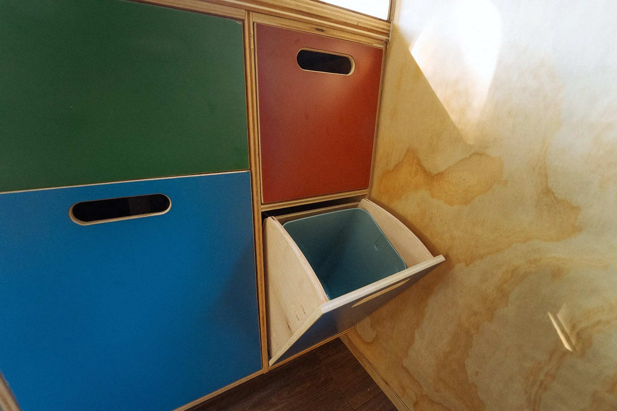 Colorful cubby storage unit with blue, green, and red compartments, and an open foldable cover revealing a blue storage bin inside, against a yellow marbled wall.