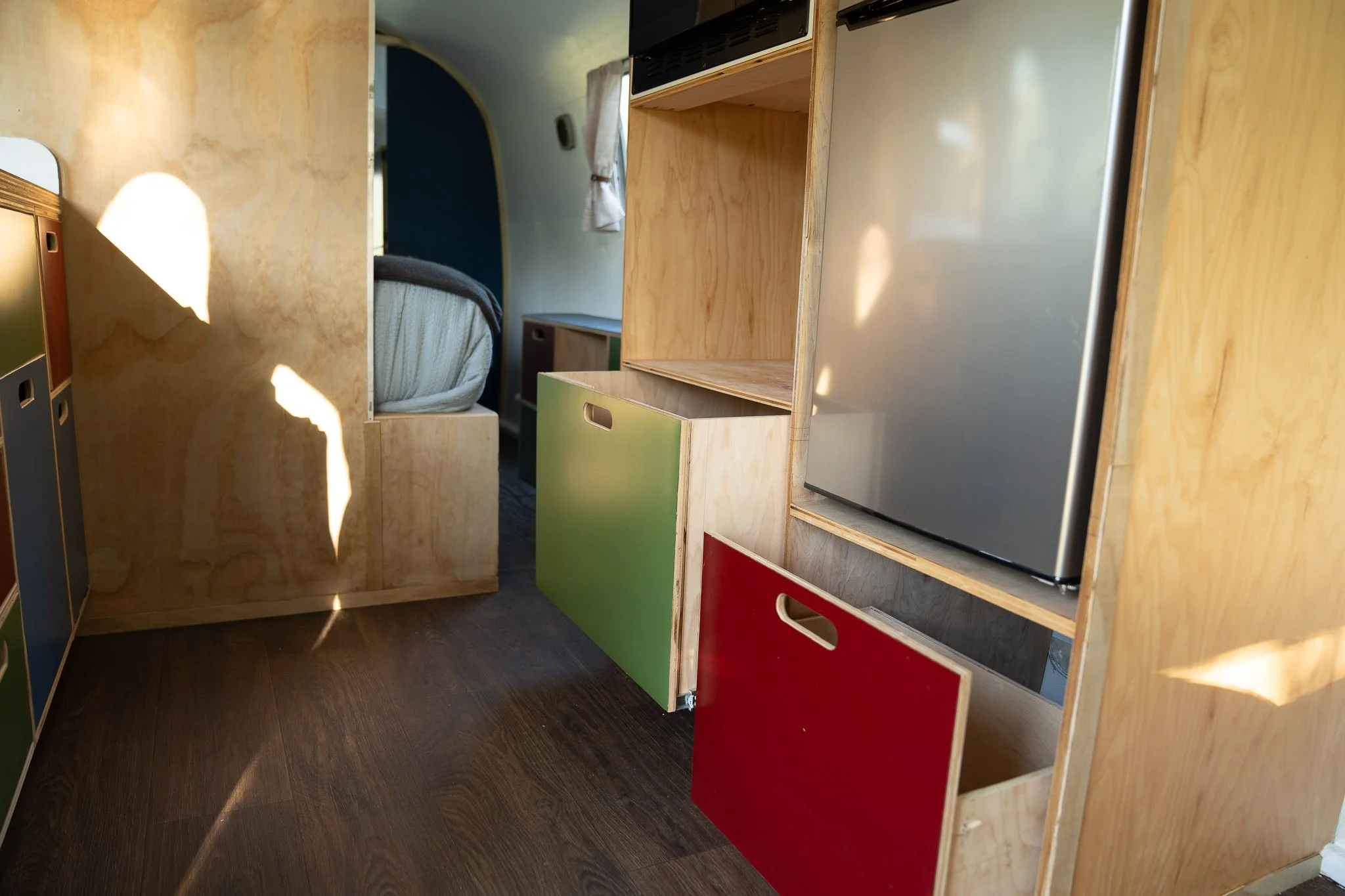 Interior of a tiny home or camper with colorful storage cabinets, a stainless steel fridge, and a view into a sleeping area with a bed.