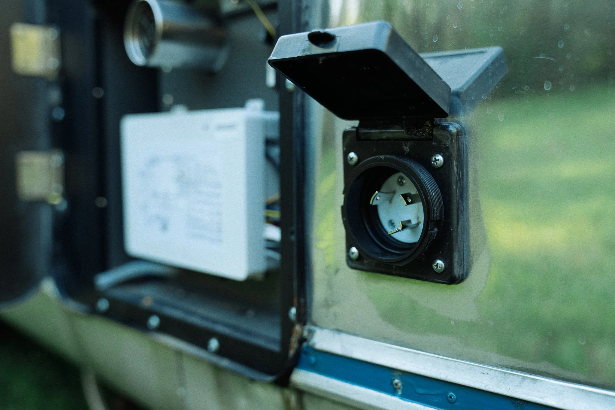Close-up of an outdoor power outlet with a protective cover on a metal surface, with electrical wiring and a white electrical component in the background.