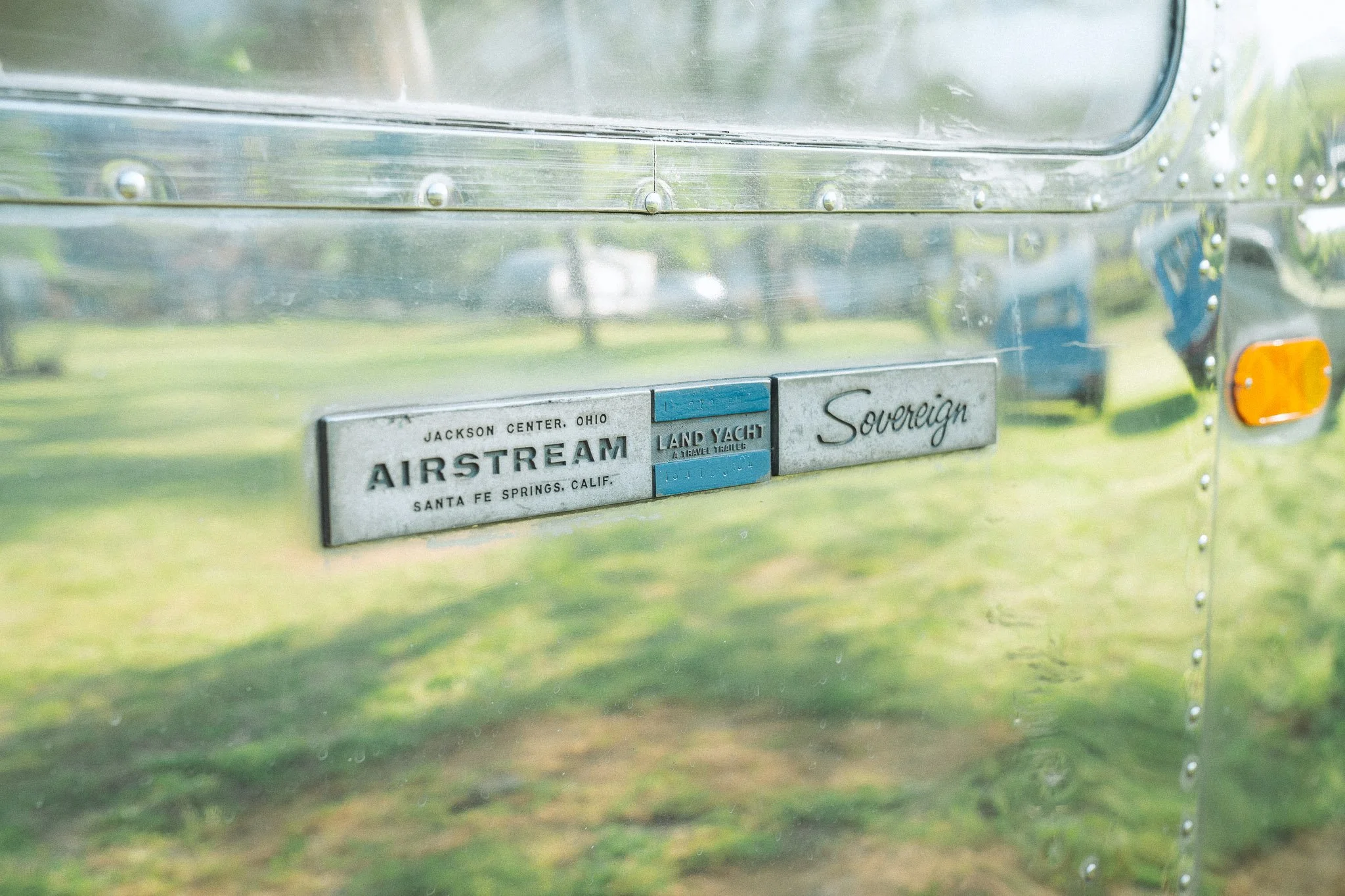 Close-up of a vintage silver Airstream trailer with brand and model badges, including 'Jackson Center Ohio', 'Airstream', 'Santa Fe Springs Calif.', 'Land Yacht', and 'Sovereign'; reflects the surrounding grass and trees.