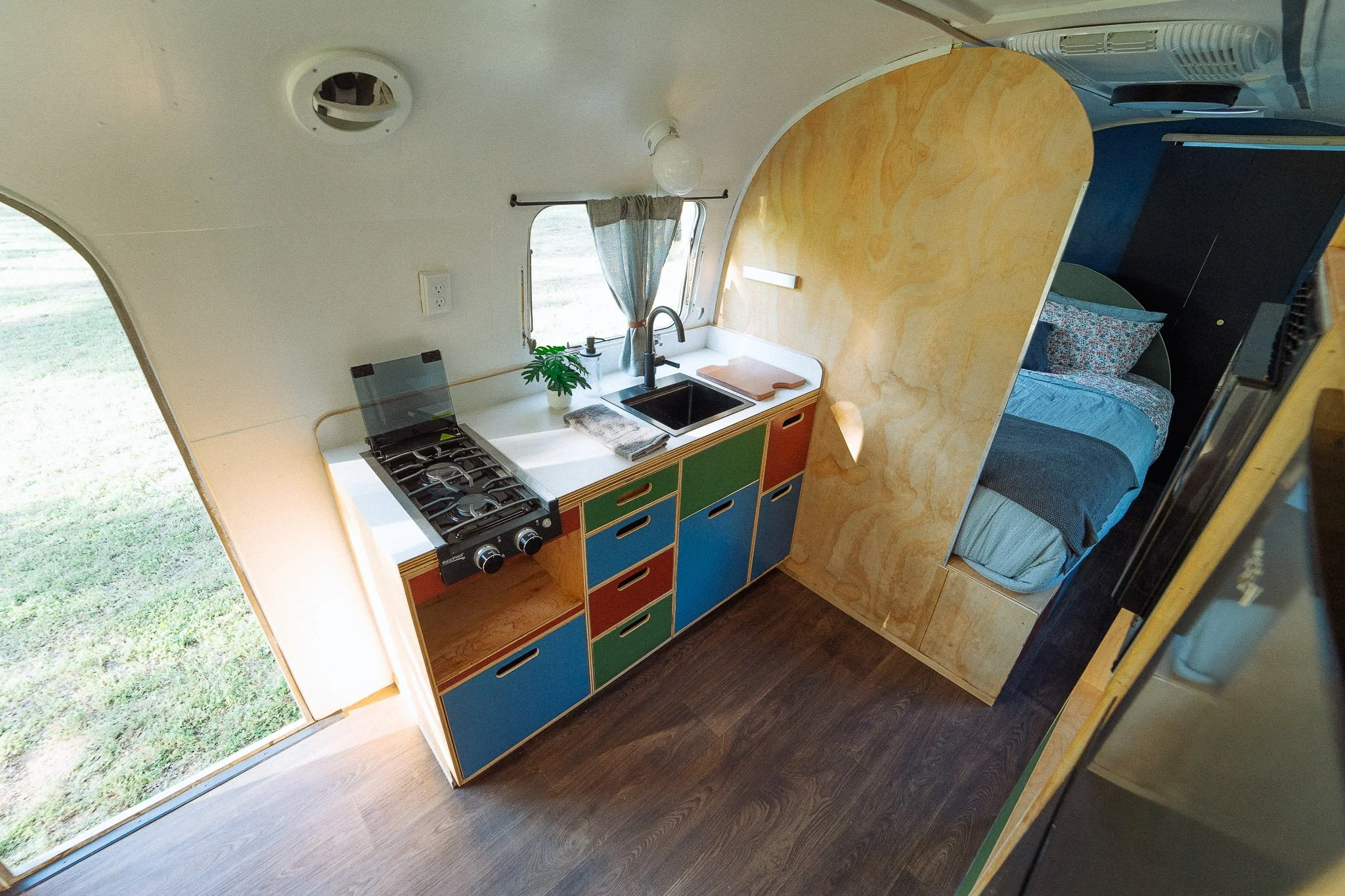 Interior of a compact living space with a kitchenette, colorful cabinets, a sink, stovetop, window with curtains, a plant, and a sleeping area with a bed and pillows.
