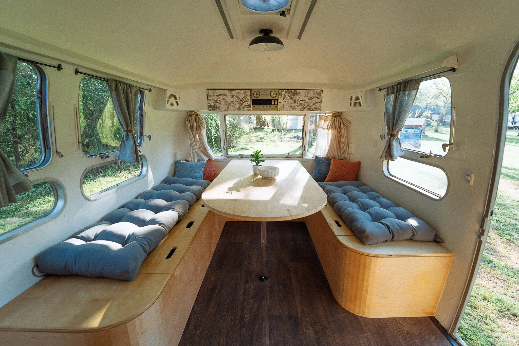 Interior of a cozy camper with built-in bench seating on both sides of a wooden table, decorated with cushions, and surrounded by windows with curtains.