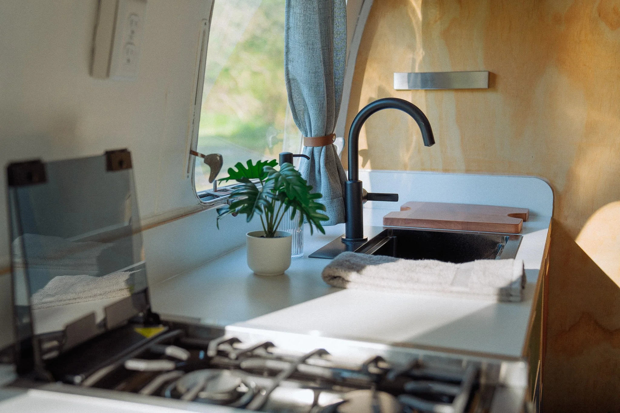A compact kitchen with a stove, a small potted plant, a window, a faucet, and some shelves with cutting boards.