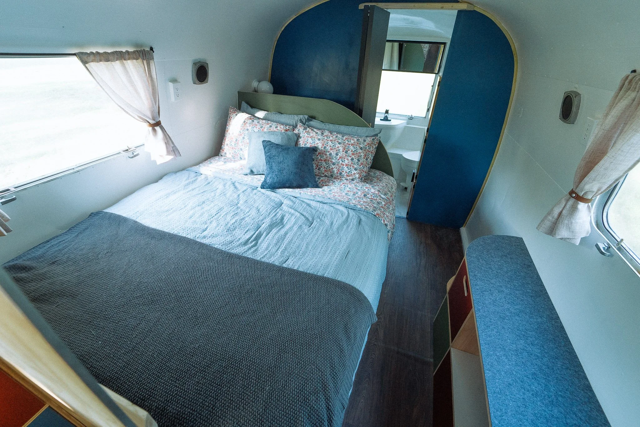 Interior of a cozy camper bedroom with a neatly made bed, small windows with curtains, and an adjoining bathroom visible through an open door.