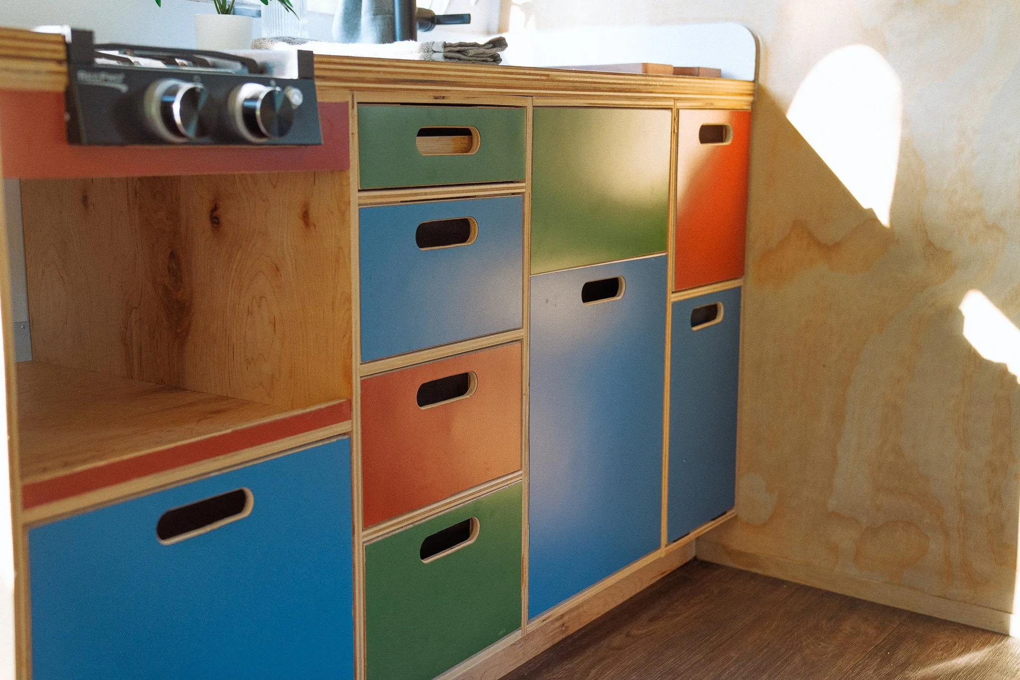 Colorful wooden storage cabinet with variously colored drawers in orange, blue, green, and natural wood finish, placed against a yellow wall with sunlight streaming in.