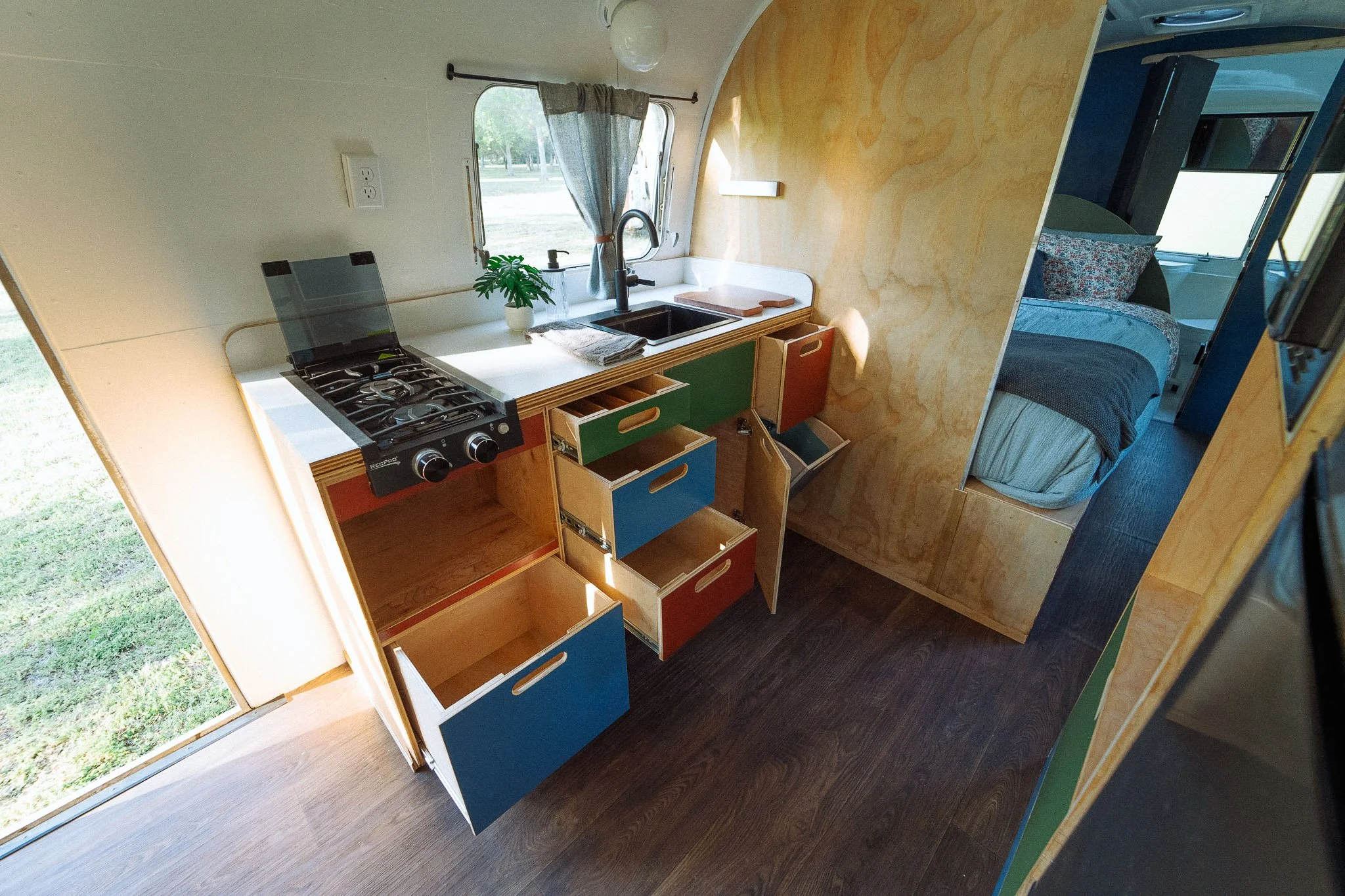 Interior view of a tiny home or camper with a small kitchen area featuring a stove, a sink, and colorful drawers, alongside a bedroom visible through a partial wall.