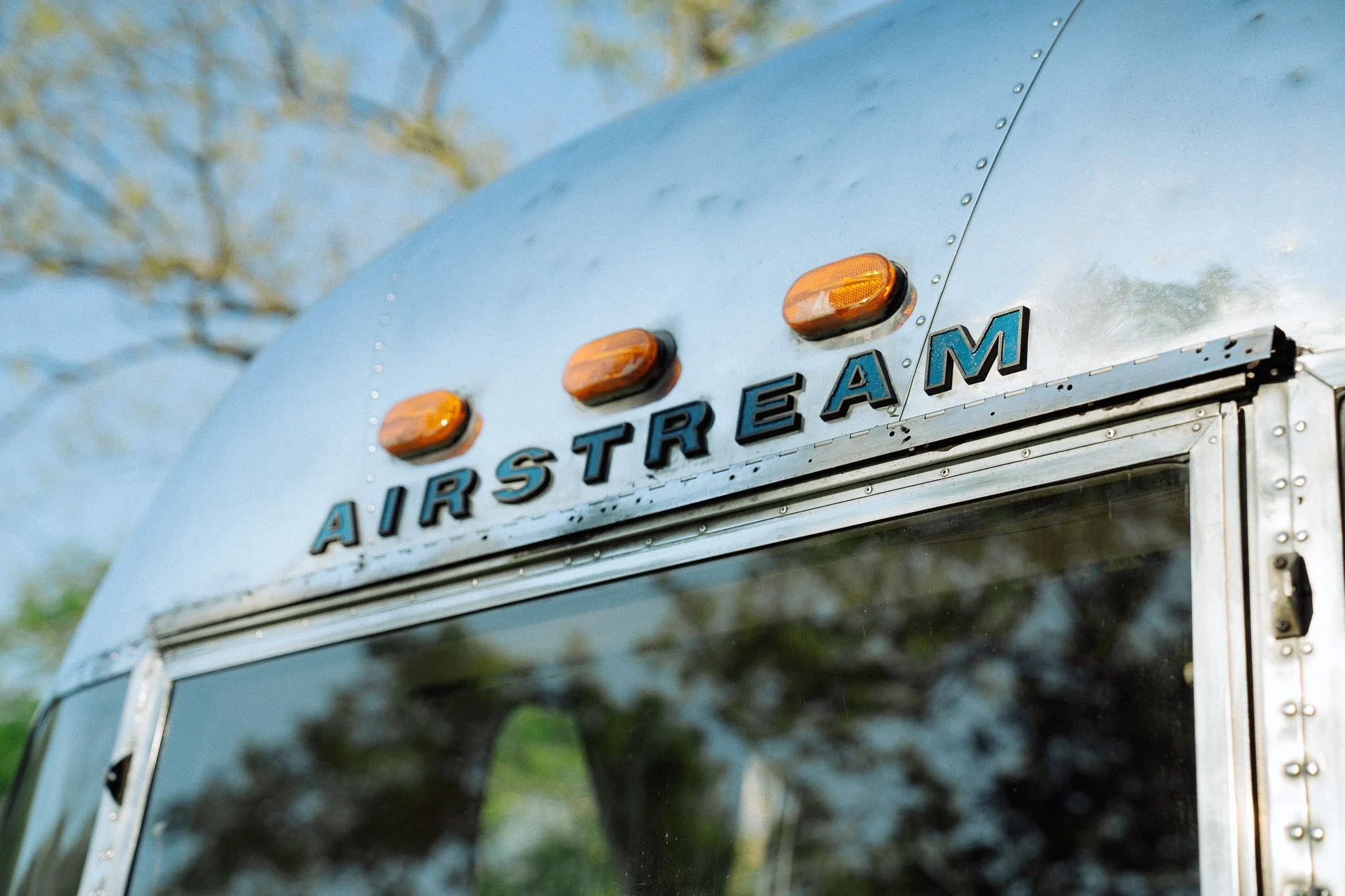 Close-up of the front of an Airstream trailer with the brand name in black letters, three amber lights above, and a reflective windshield, with trees in the background.