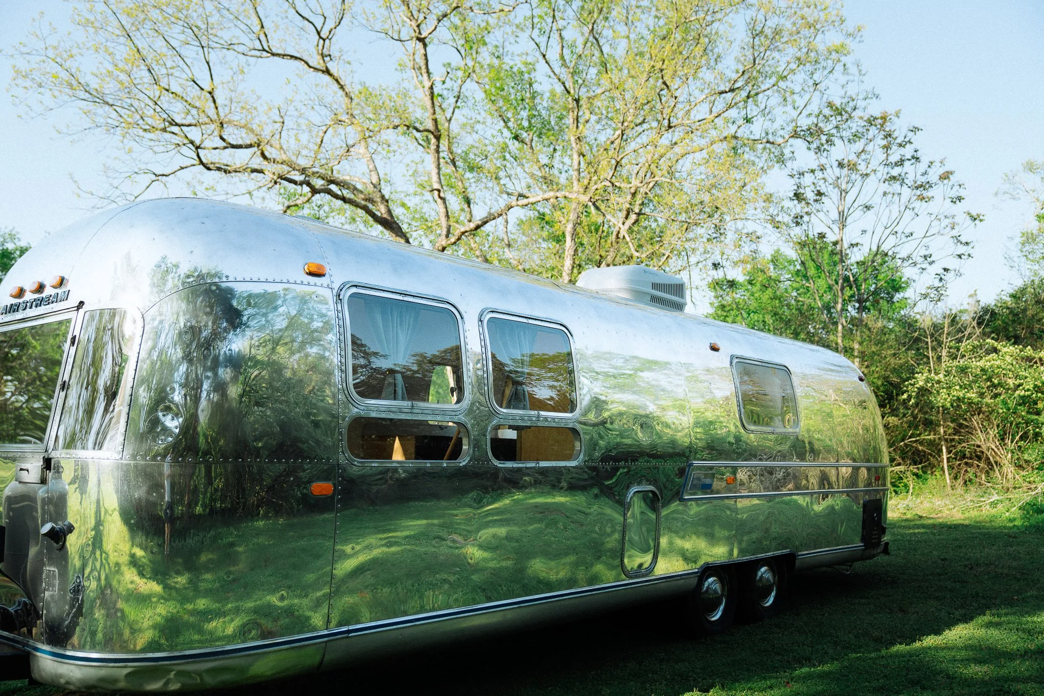 A shiny, silver travel trailer parked on a grassy area with trees in the background.