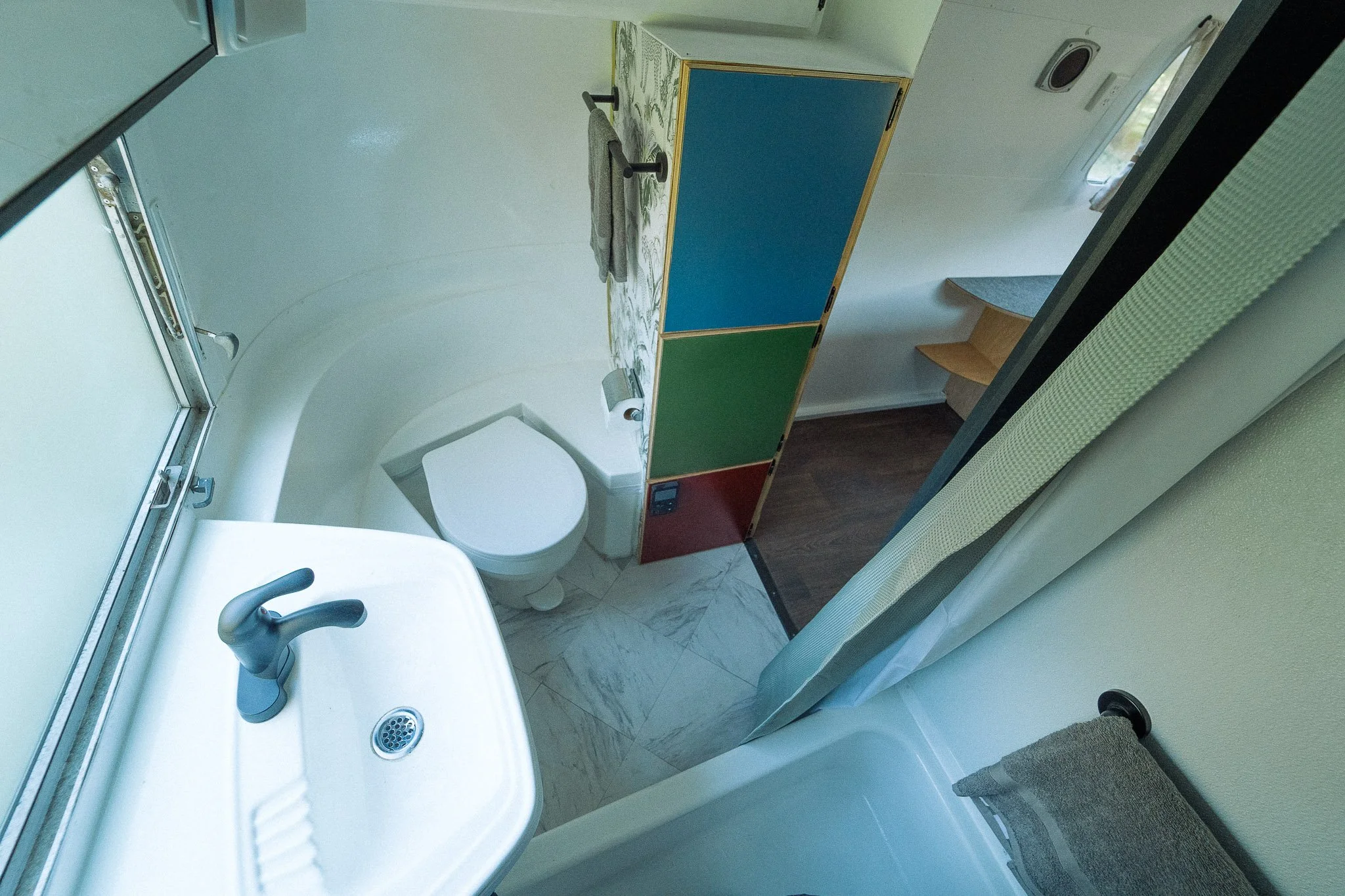 Small bathroom with a white toilet, a small white sink with a black faucet, a mirror, a partial shower curtain, and a colorful folding room divider with blue, green, and red sections. A window, wooden step, and small decorative rug are also visible.