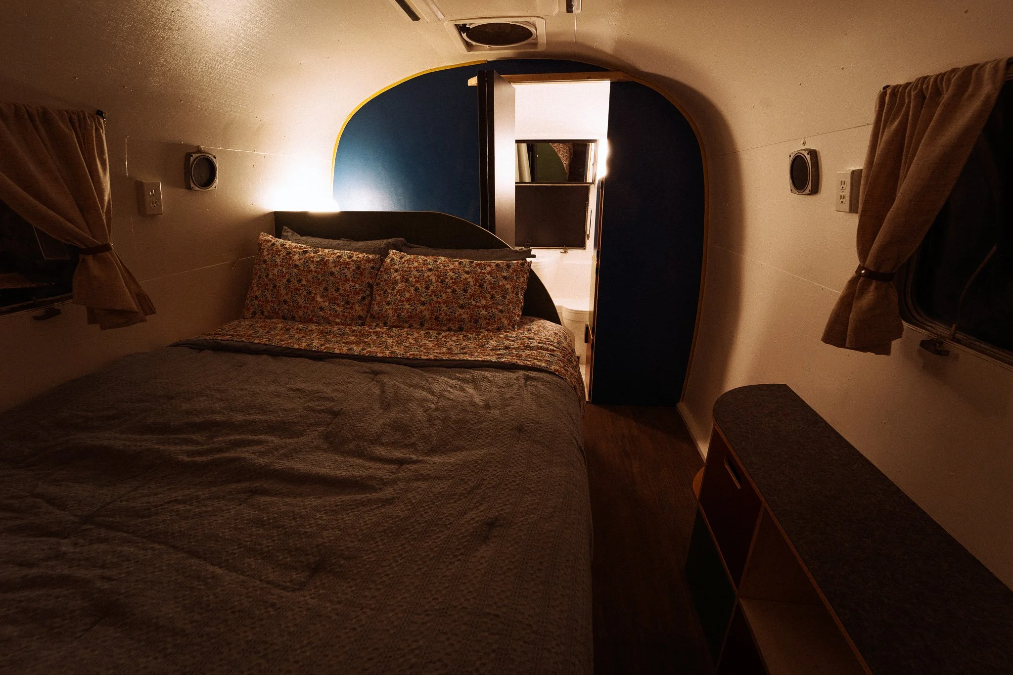 Interior of a small bedroom with a bed, floral bedding, two small windows with curtains, and a door leading to a bathroom.