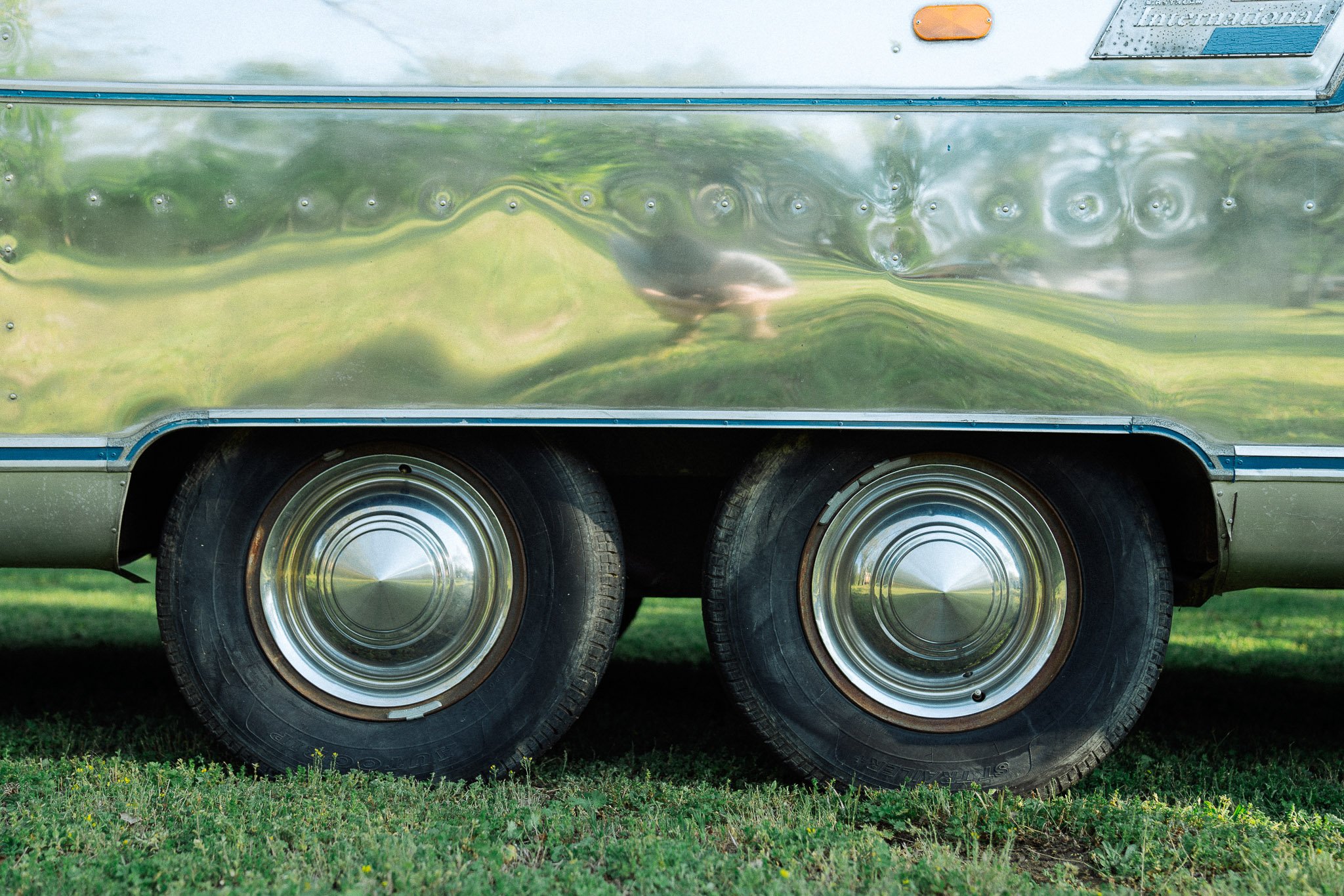 Close-up of a vintage silver trailer with two shiny hubcap wheels on green grass, with a reflection of a bird and grass on the trailer's surface.