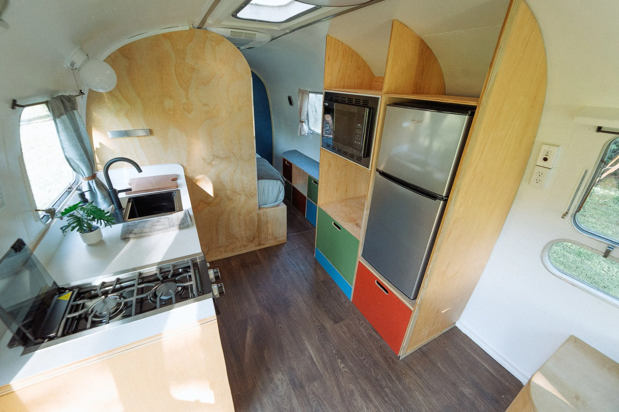 Interior of a tiny home or RV with wooden cabinetry, a refrigerator, microwave, small kitchenette with a sink and a potted plant, a bed with a window and curtains, and colorful storage drawers.