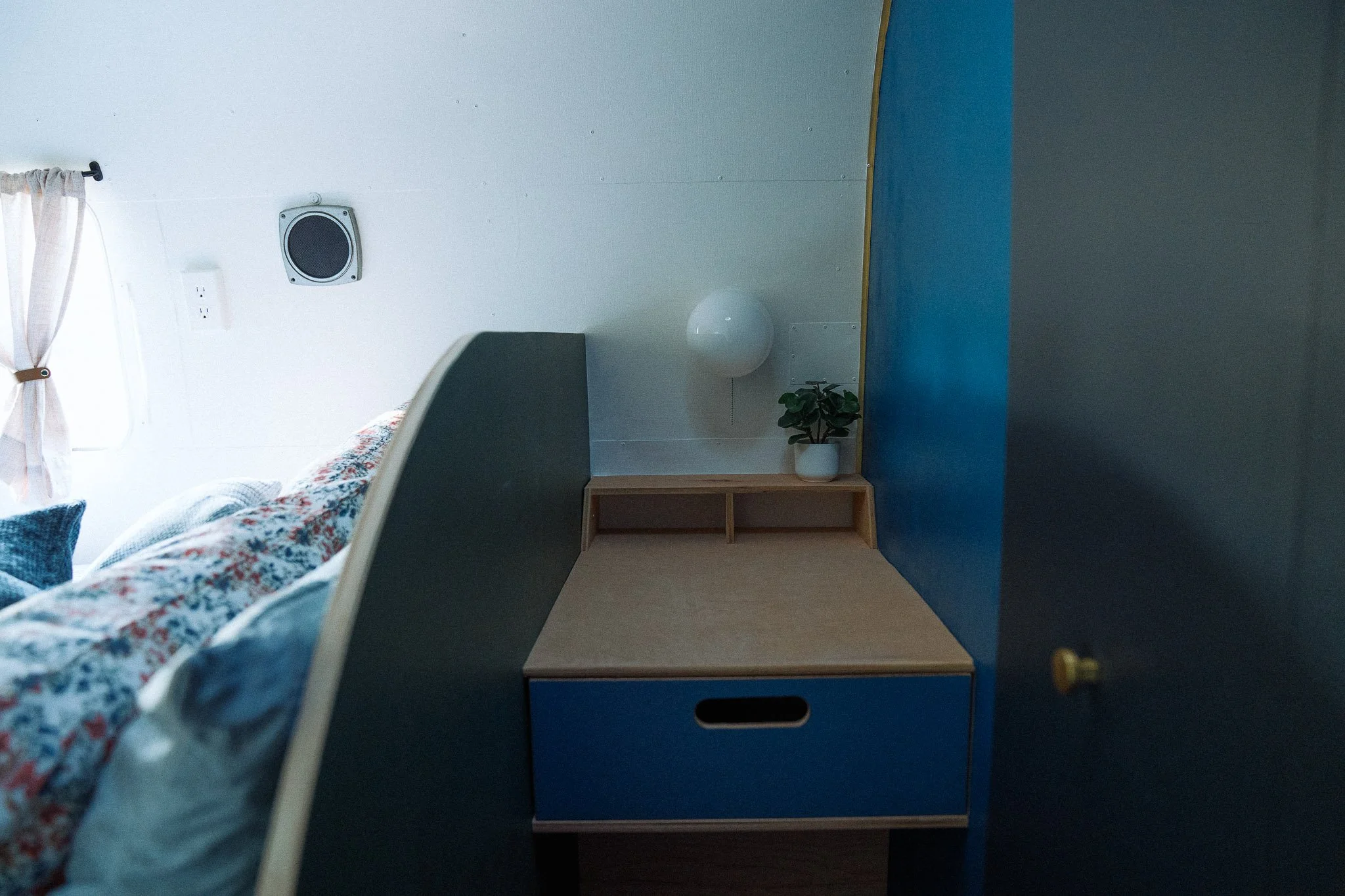 Interior of a bedroom with a bed partially visible on the left, a window with a white curtain, and a blue wall. A small wooden bedside table with a potted plant and a wall-mounted lamp is visible on the right side.