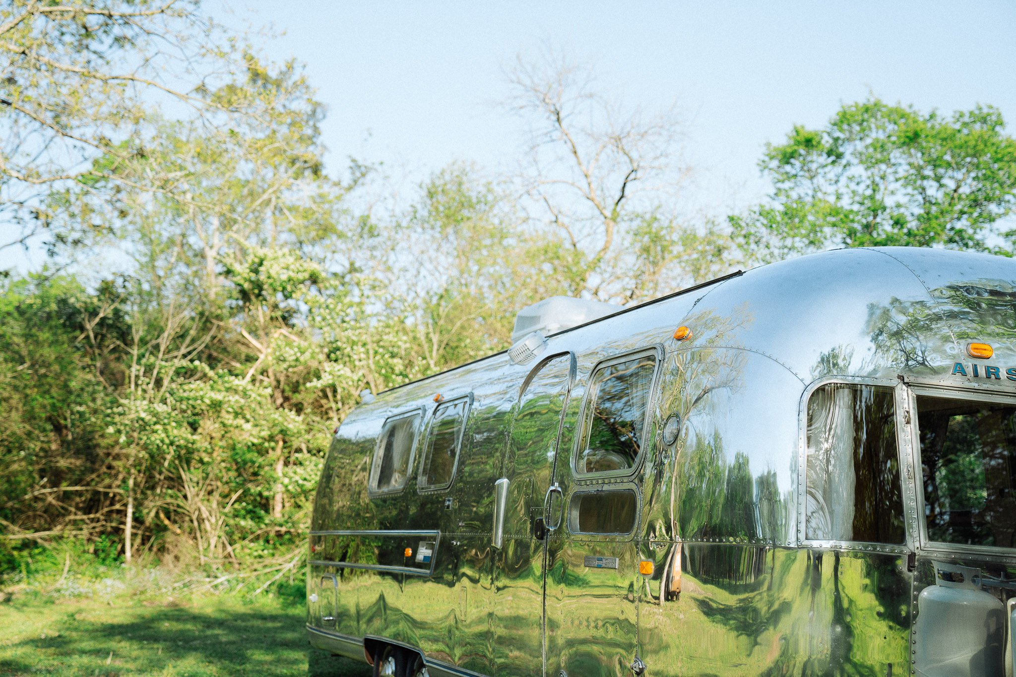 A shiny, metallic camper trailer parked on green grass with trees and a blue sky in the background.