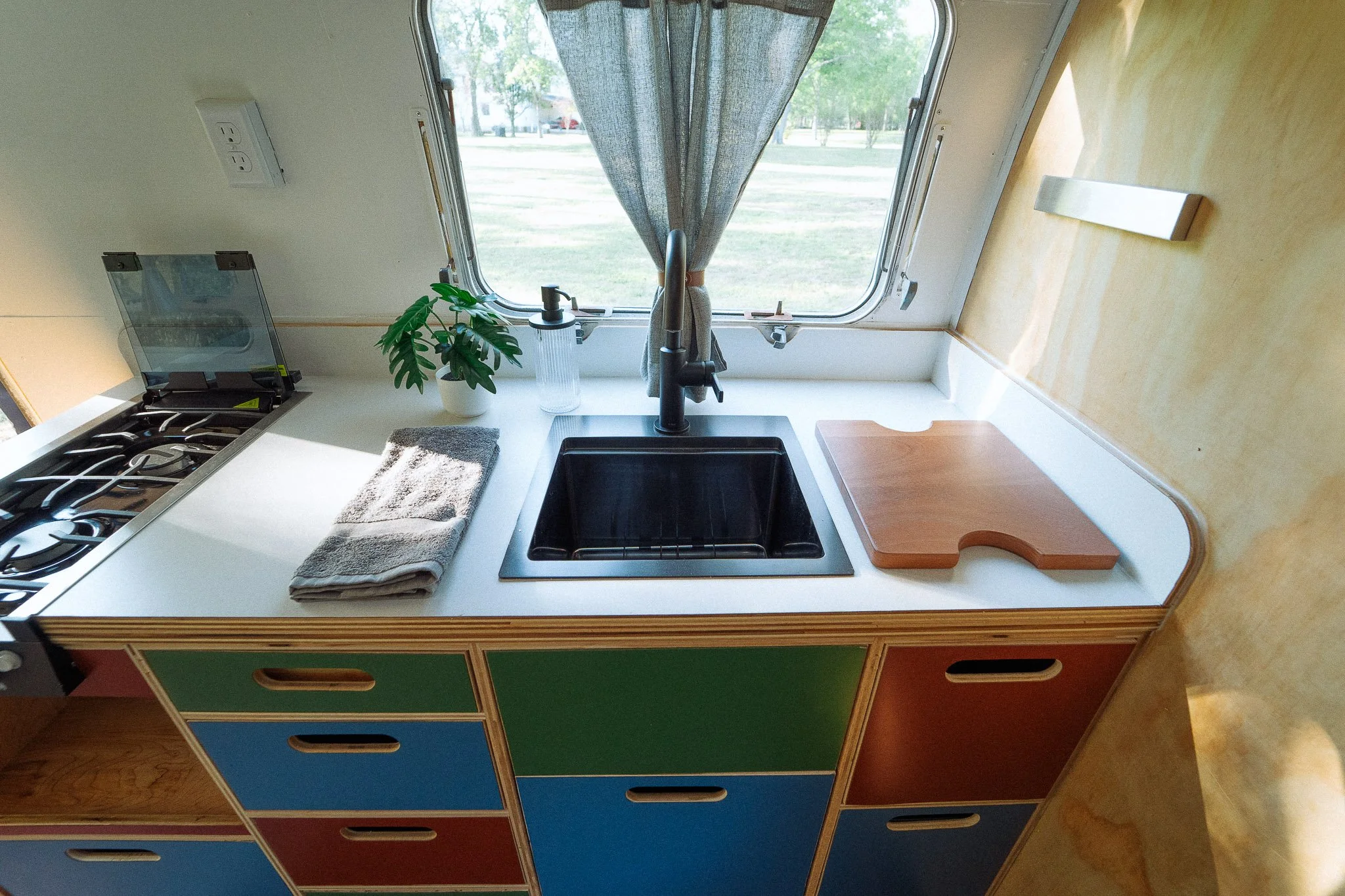 Kitchen countertop with a sink, a cutting board, a potted plant, a dish soap bottle, and a window with a curtain showing an outdoor view.