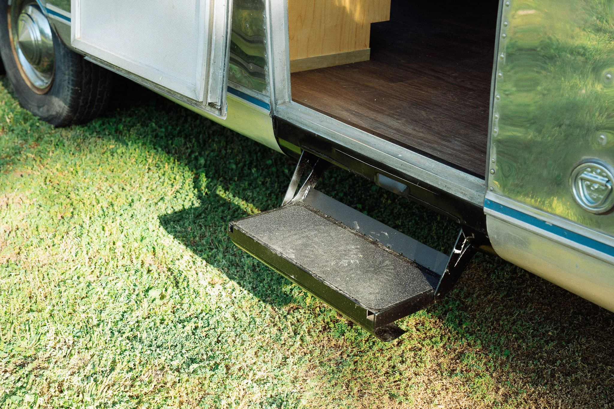 Close-up of a fold-down metal step attached to the side of a silver travel trailer, with the trailer open to reveal a wooden interior floor, parked on green grass.