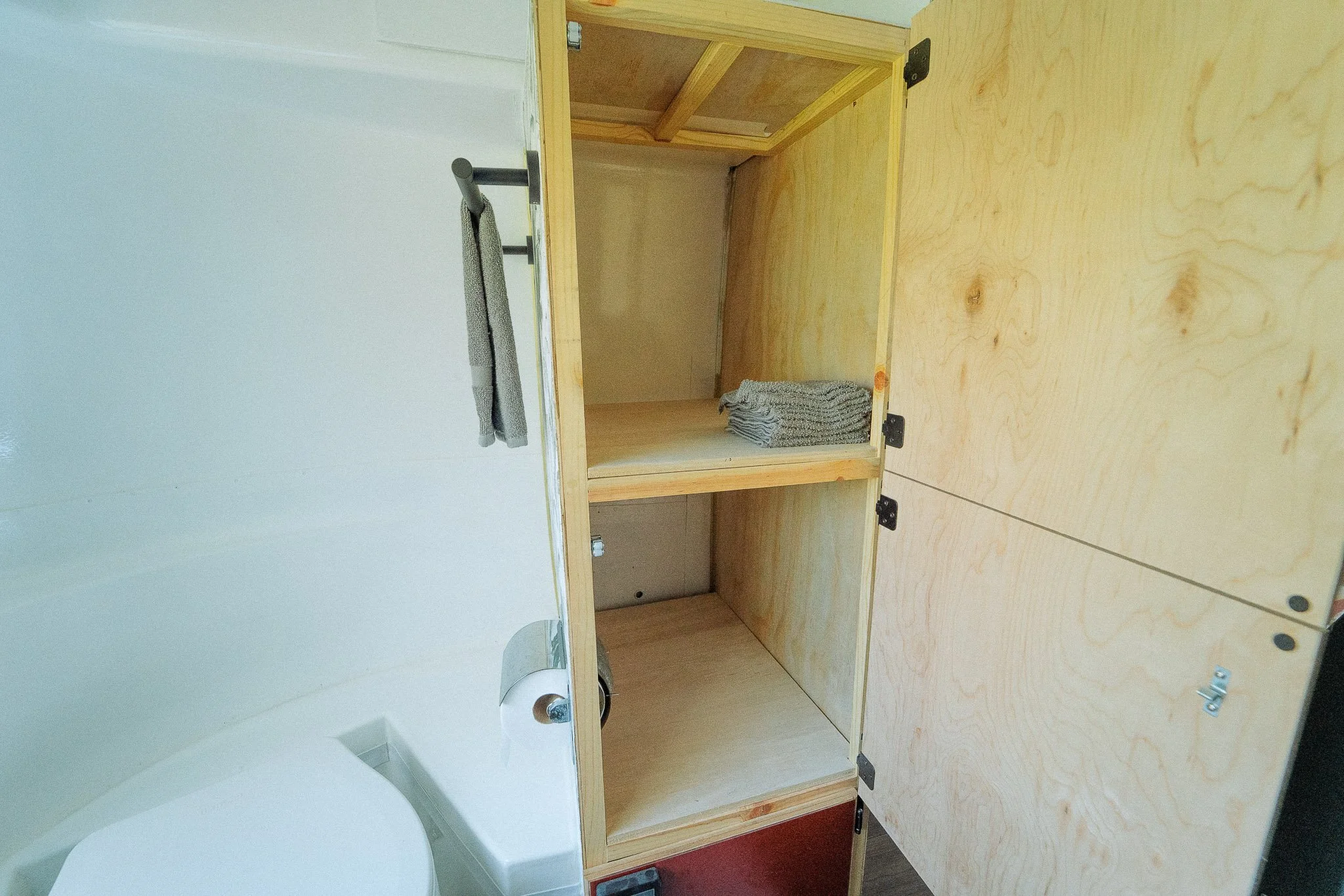 An unfinished wooden cabinet with open shelves, holding folded gray towels, next to a white bathtub.