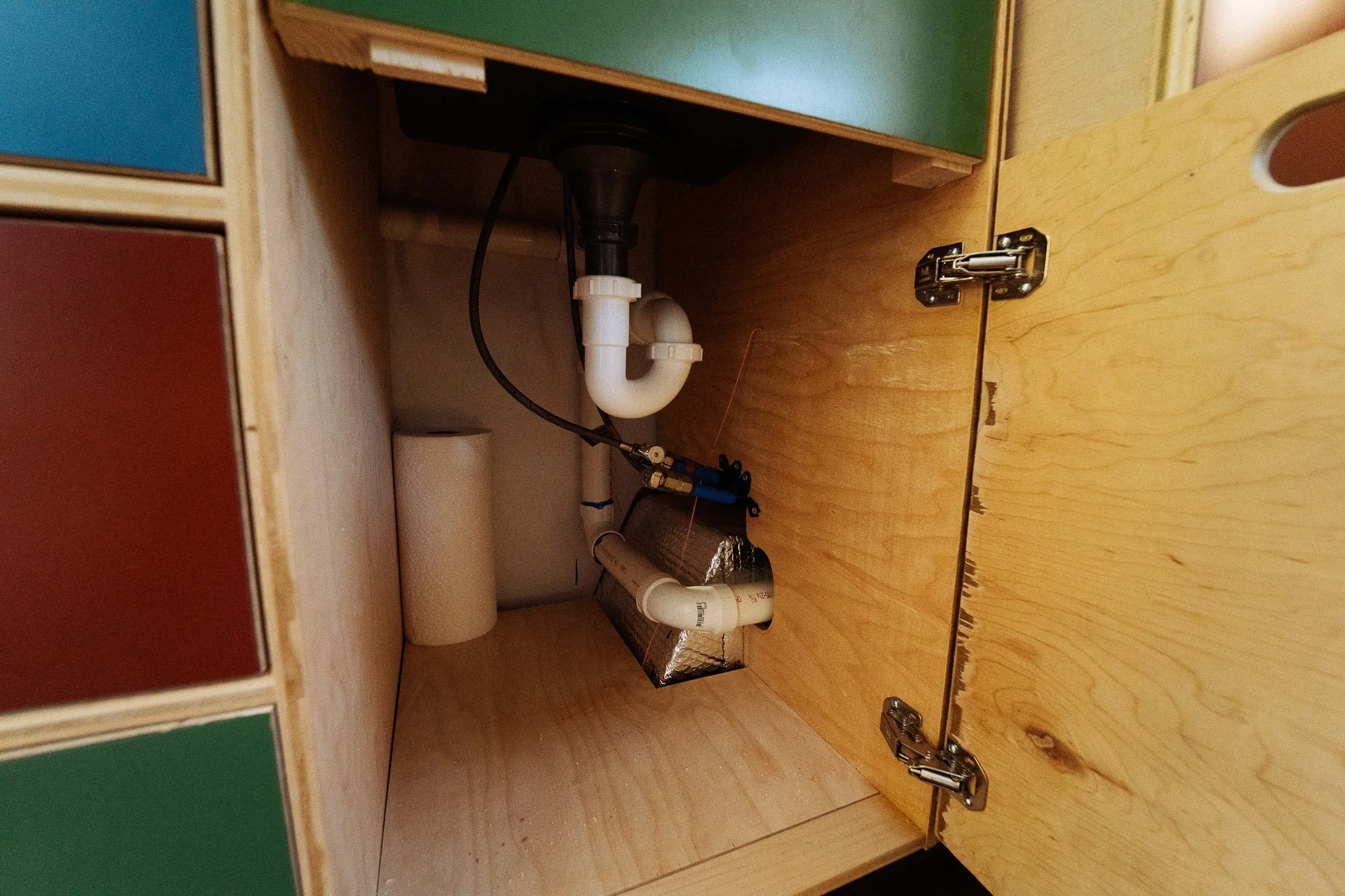The image shows the inside of a wooden cabinet under a sink, with visible plumbing pipes and a roll of paper towels.