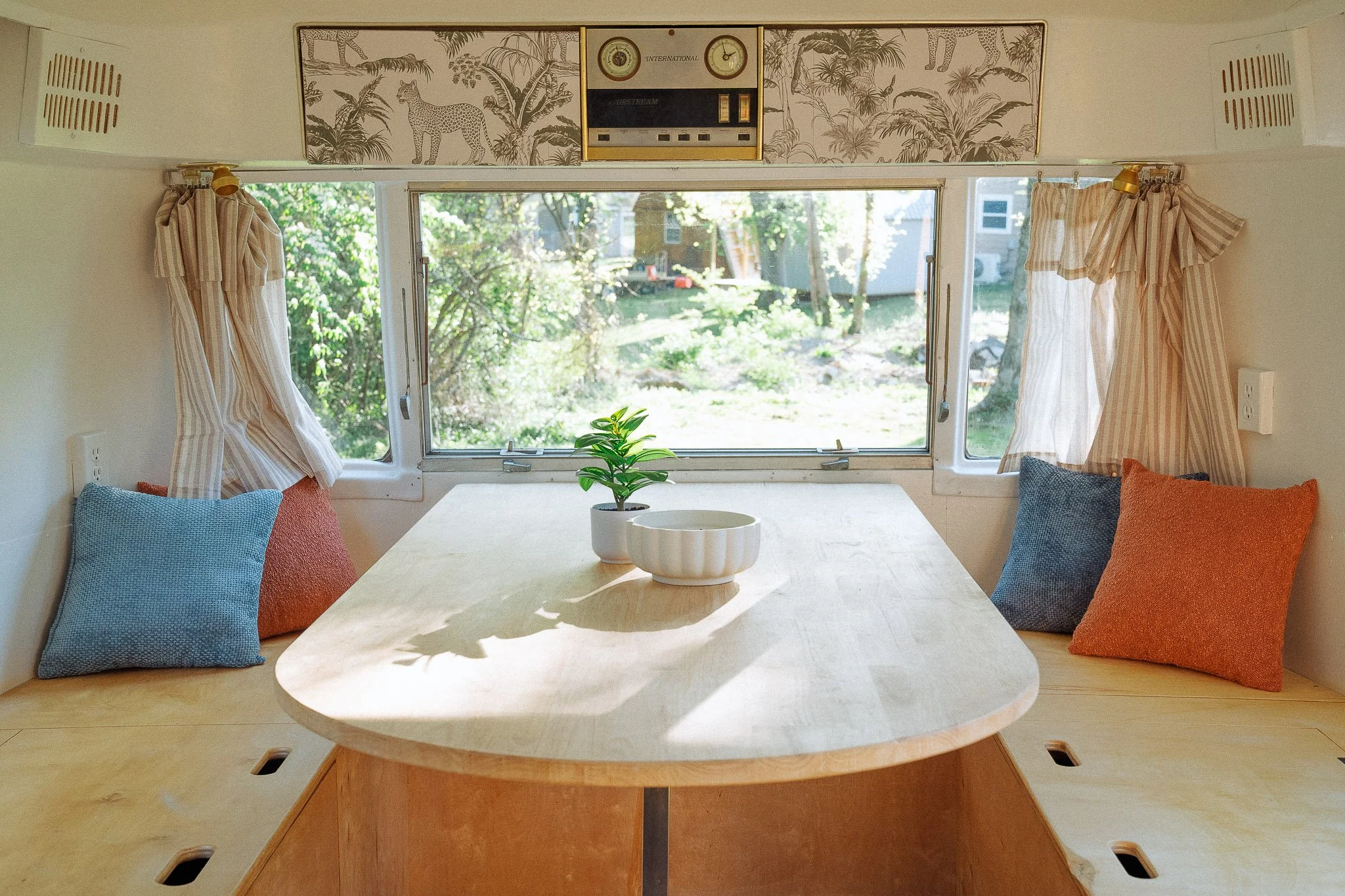 A cozy dining nook with a wooden table, a potted plant, and a bowl. Behind the table, a large window shows a green backyard. Cushions in blue and orange are on built-in benches along the walls, with striped curtains on either side of the window. The 