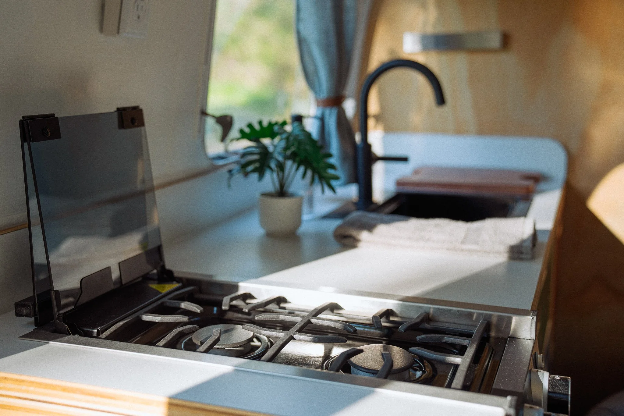 Kitchen sink with black faucet, a small potted plant, a window with curtains, and a stove in a compact kitchen.