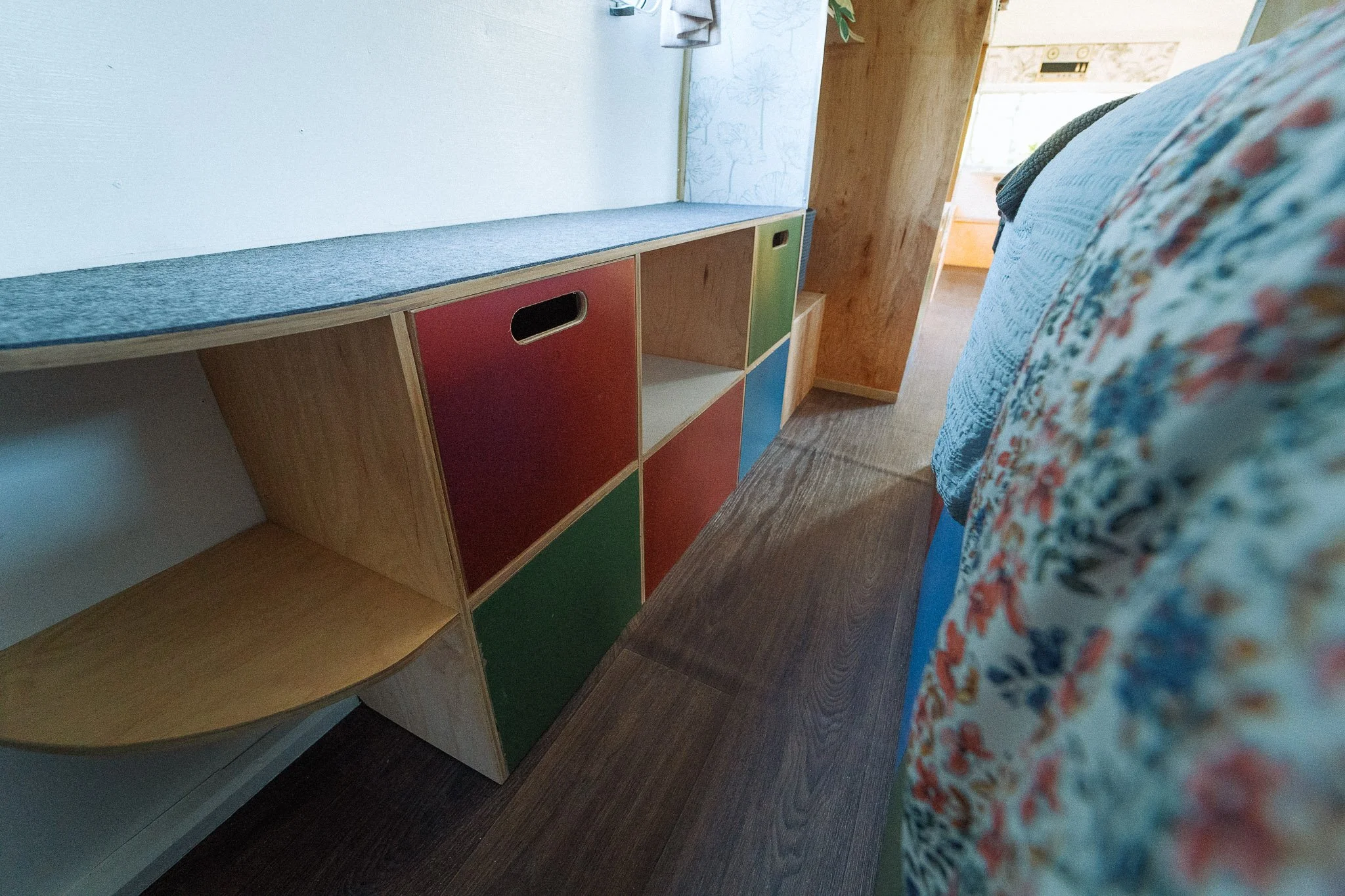A wooden storage unit with color-blocked doors in green, red, and blue, situated beside a bed with floral bedding, in a cozy room with wooden paneling and a hint of a window in the background.