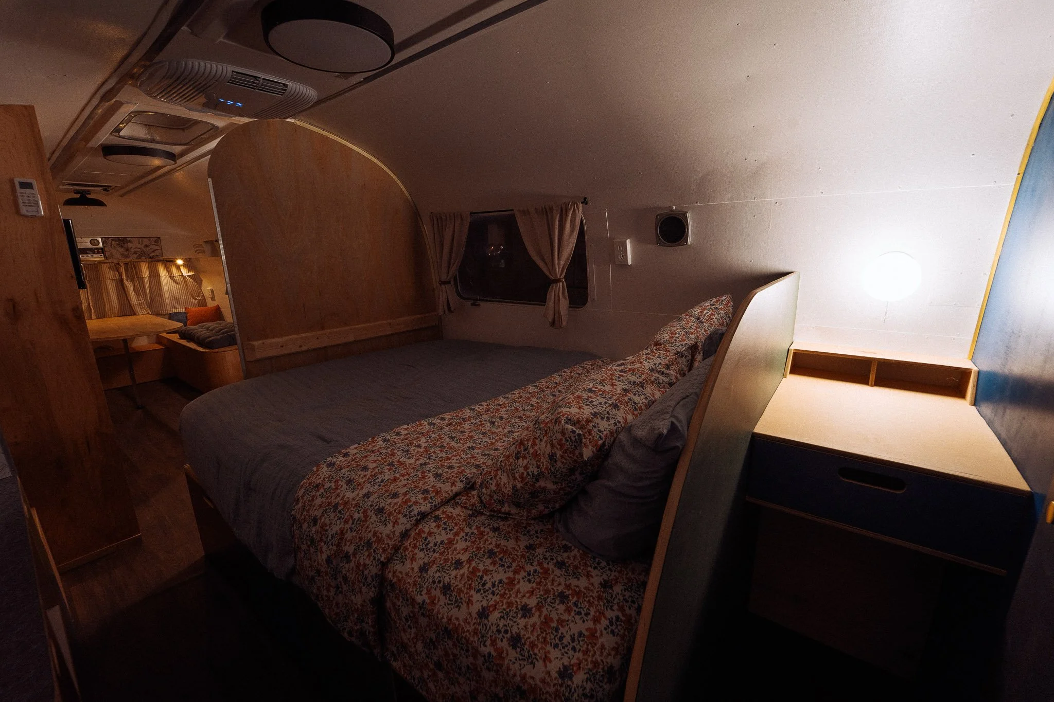 Interior of a cozy bedroom with a bed covered in a floral and dark blanket, a window with beige curtains, and a small side table with a light on. There appears to be a sitting area with cushions and curtains in the background.