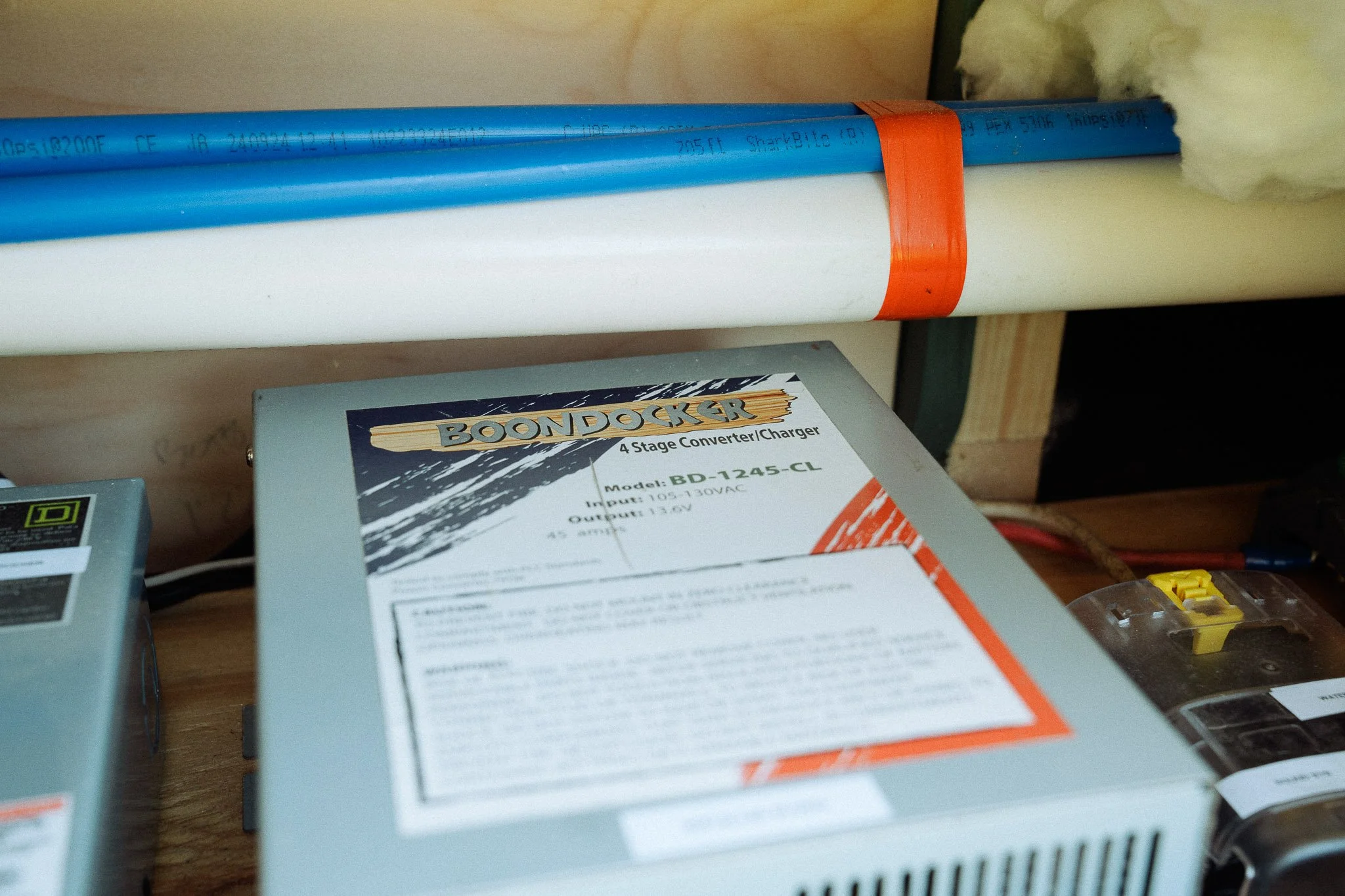 Close-up of a gray Boondocker 4-stage converter/charger with model BD-1245-CL, placed on a wooden surface. Behind it, there are blue and white pipes secured with an orange strap, and to the right, there is a clear plastic circuit breaker with yellow 