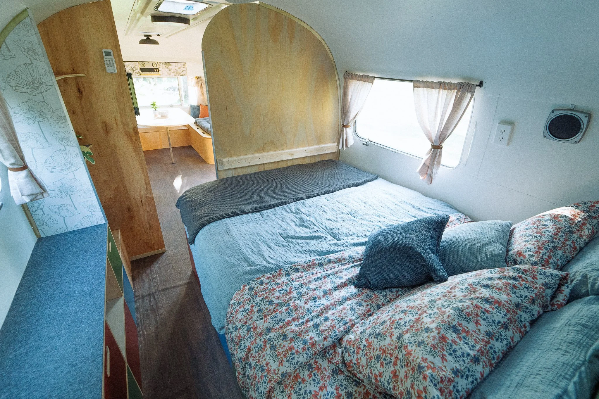 A bedroom in a camper or tiny home with a bed, pillows, and curtains on a window, with a small dining area visible through an arched wooden partition.