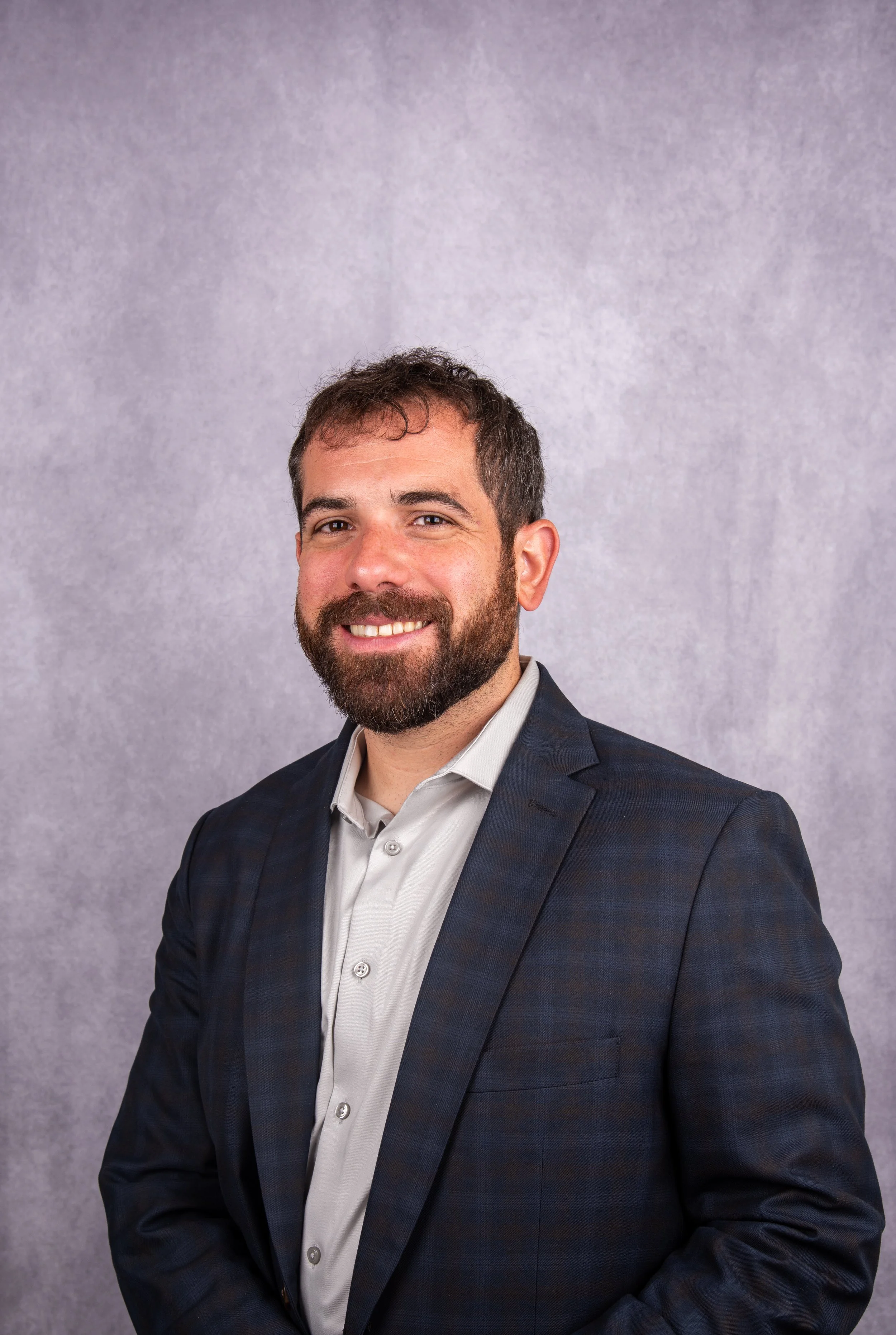 A Caucasian man with dark hair, a beard, wearing a dark blue checked suit and a white shirt, smiling against a textured light gray background.