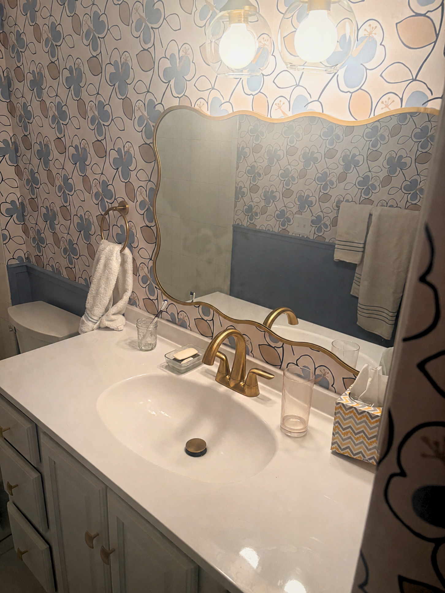 Bathroom vanity with a white countertop, gold faucet, and a mirror. On the countertop are a soap dish, an empty glass, a pink plastic cup, tissues, and a jewelry tray. There are white towels hanging on a gold towel ring, a towel rack, and a wall with