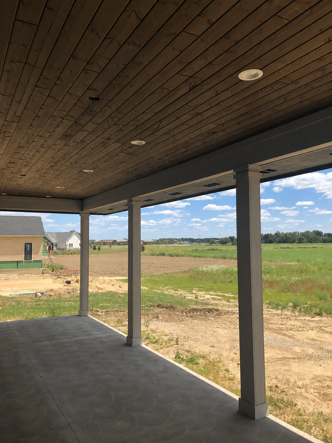 View from a porch showing a construction site with a partially built house, open fields, and a blue sky with clouds.