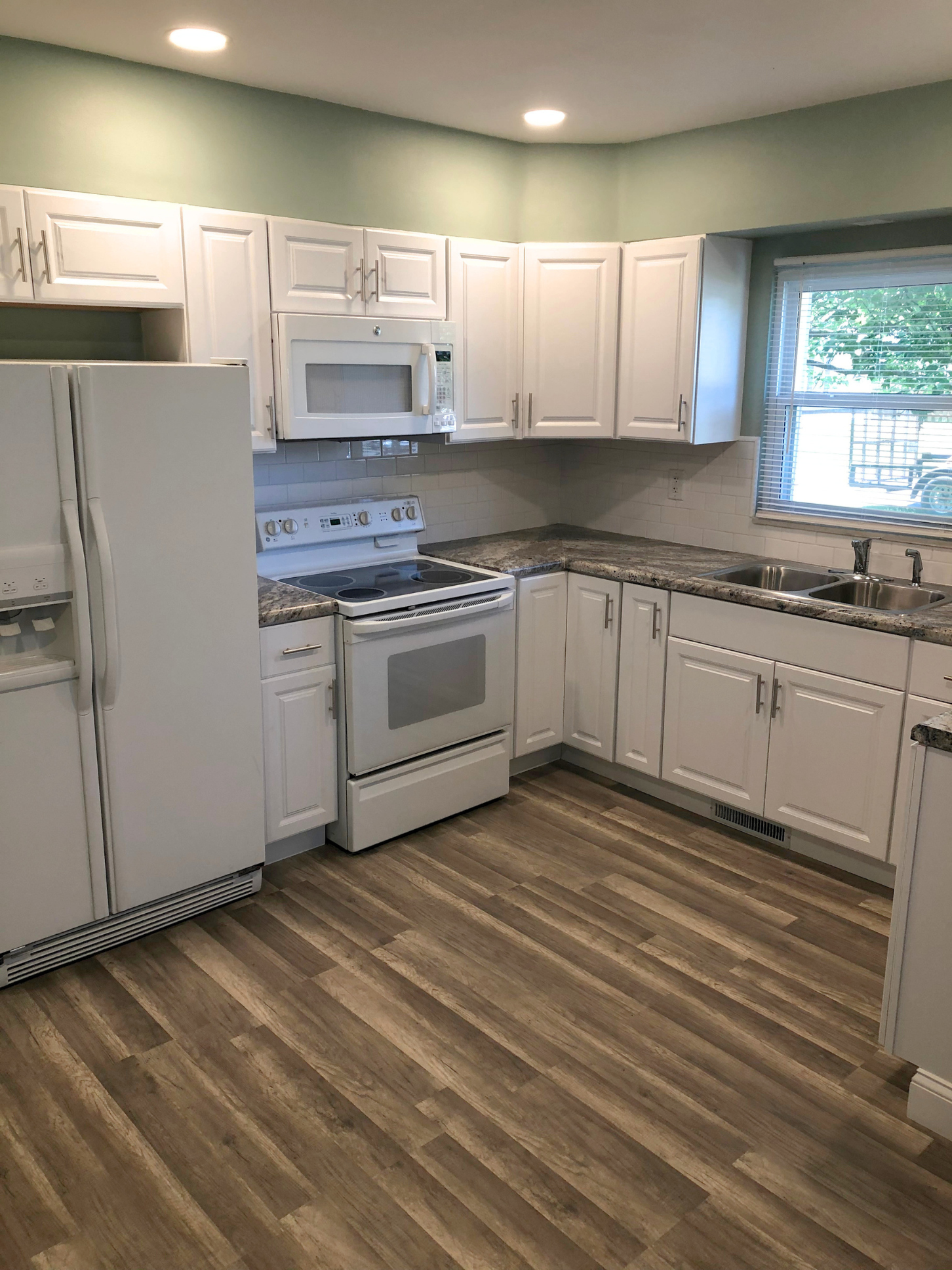 Kitchen with white cabinets, a stove, microwave, refrigerator, double sink, and wood-like flooring, next to a window with blinds.