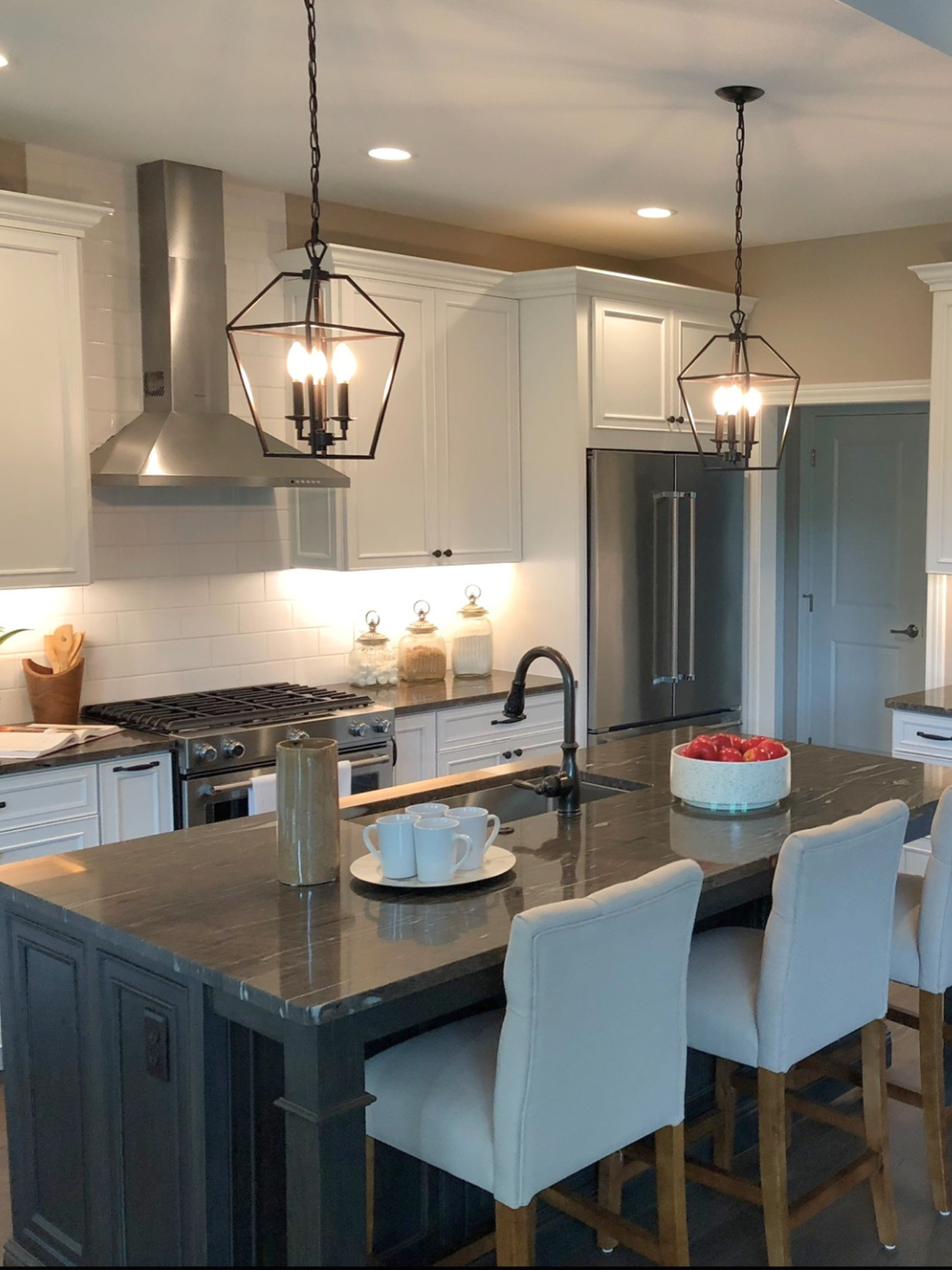 A modern kitchen with white cabinetry, a large dark island with seating, pendant lights, stainless steel appliances, and a white tile backsplash. The counter has a bowl of red apples, white mugs, and jars.