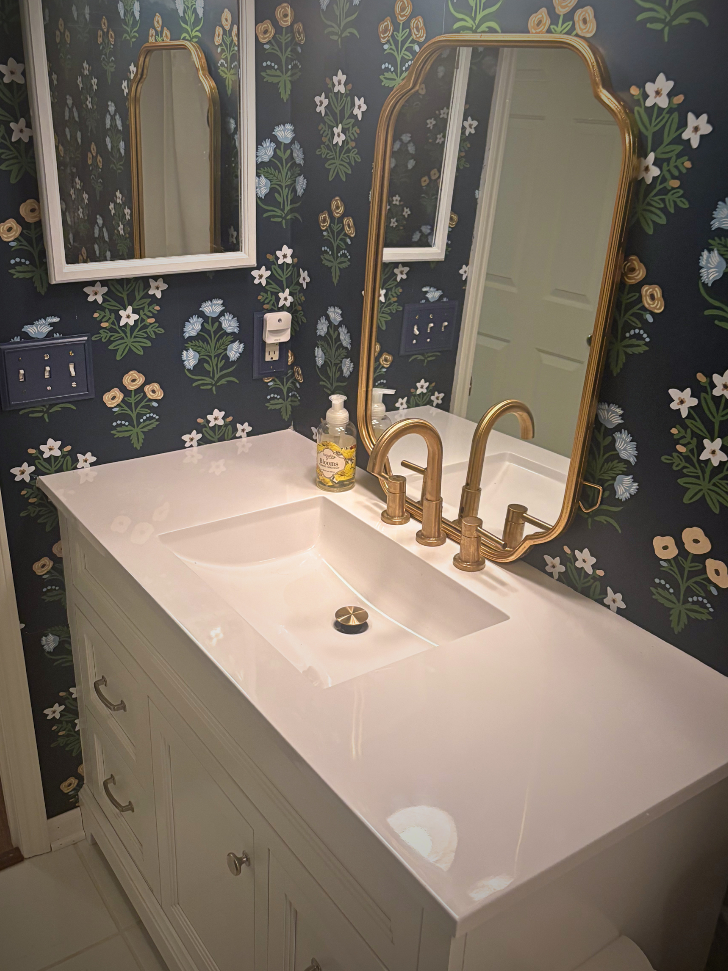 White bathroom vanity with gold fixtures and a large mirror, set against a dark floral wallpapered wall.