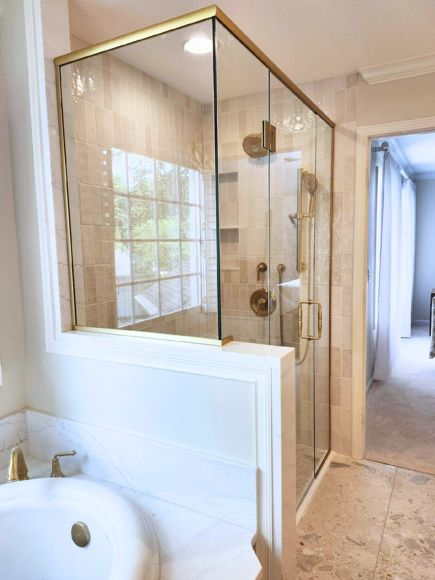 Bathroom with a glass shower enclosure, beige tile walls, a built-in shower shelf, and a window reflecting trees outside. Part of a bathtub with a gold-colored faucet is visible in the foreground.