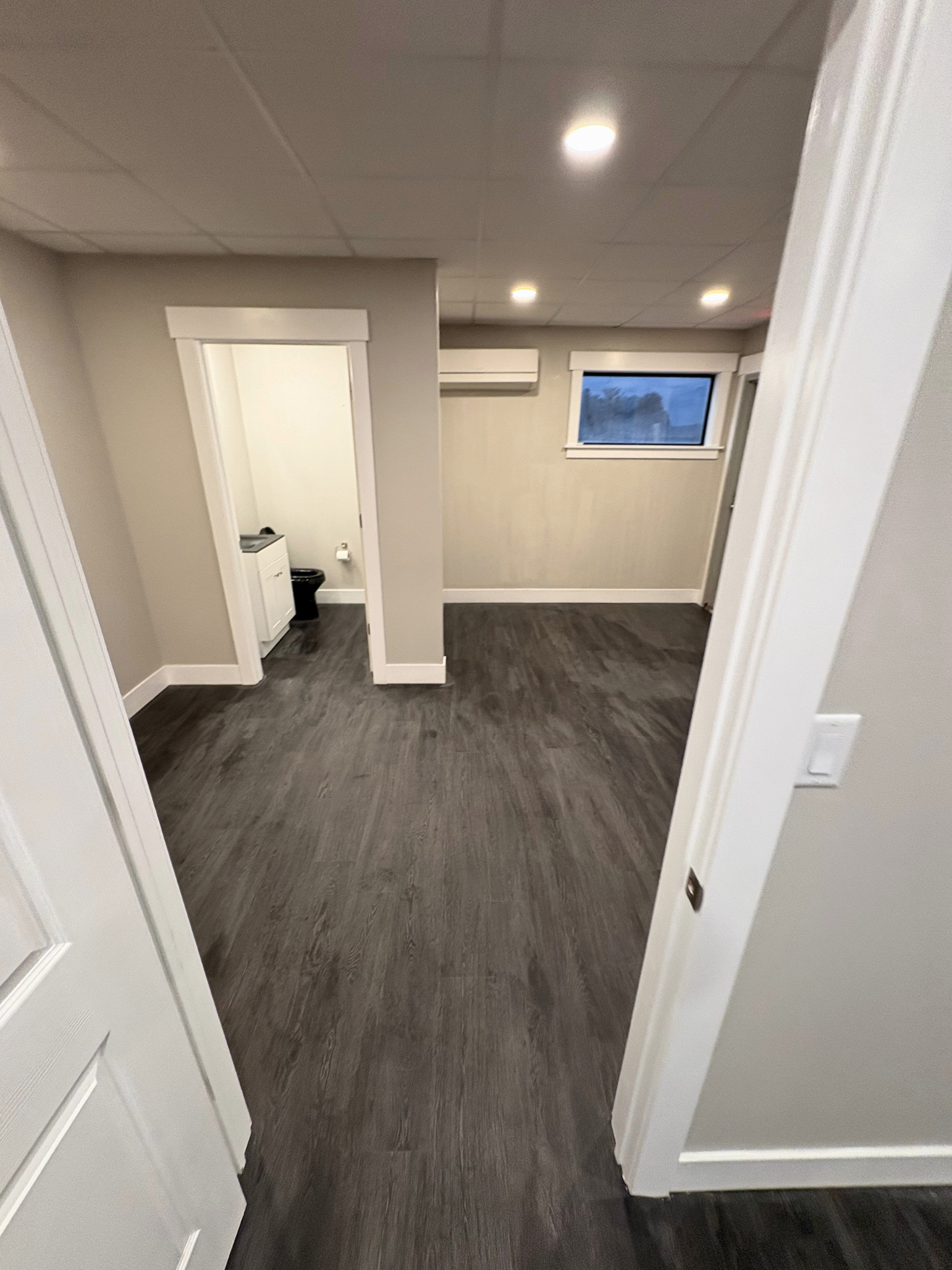 Empty room with dark wood floors, light-colored walls, small window, and ceiling lights, viewed from doorway.