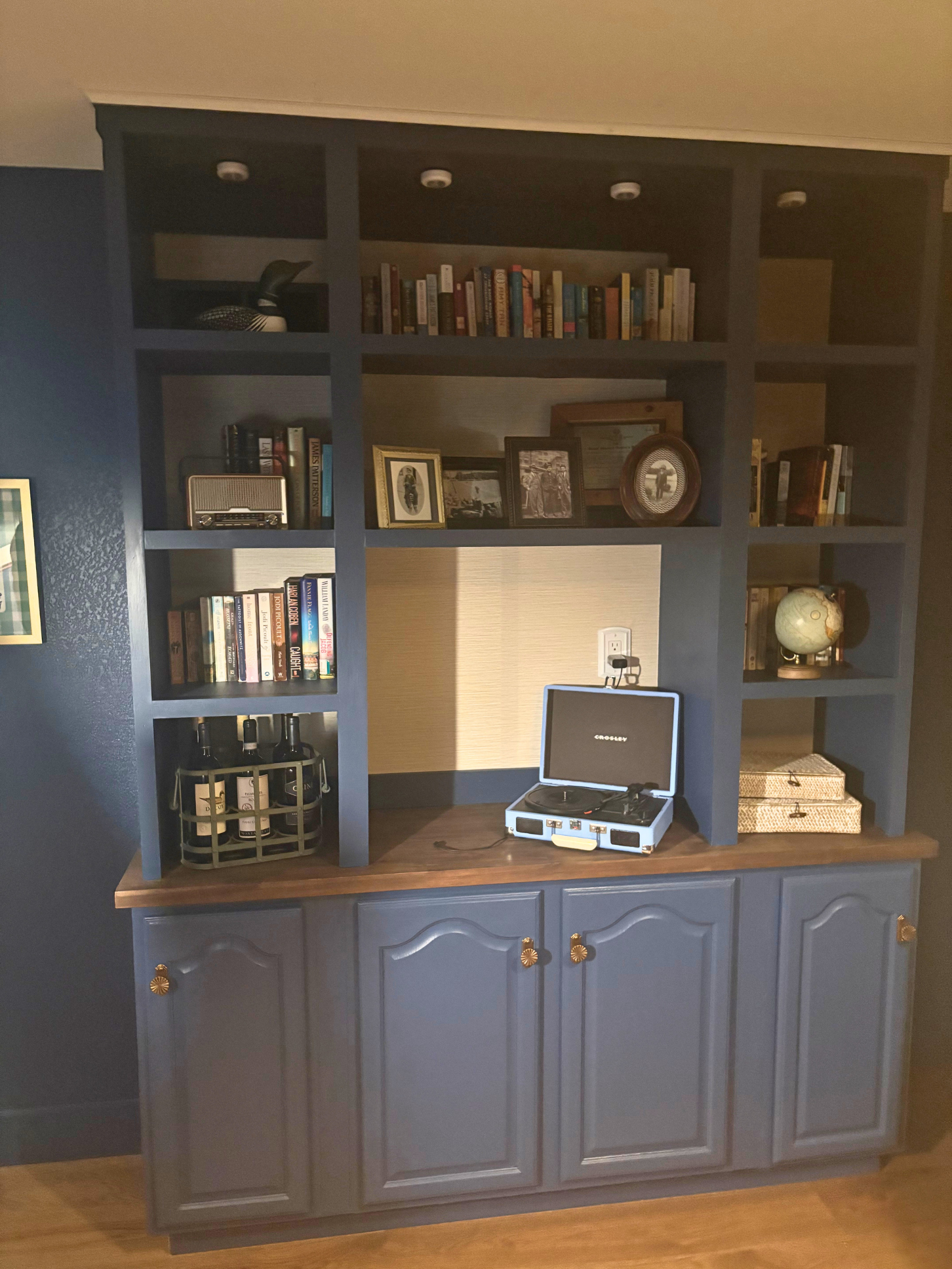 A blue wooden cabinet with multiple open shelves and a closed lower section with gold knobs. The top shelf holds a black and white rubber duck and a few books. The middle shelves display framed photographs, artwork, and a small world globe. The lower