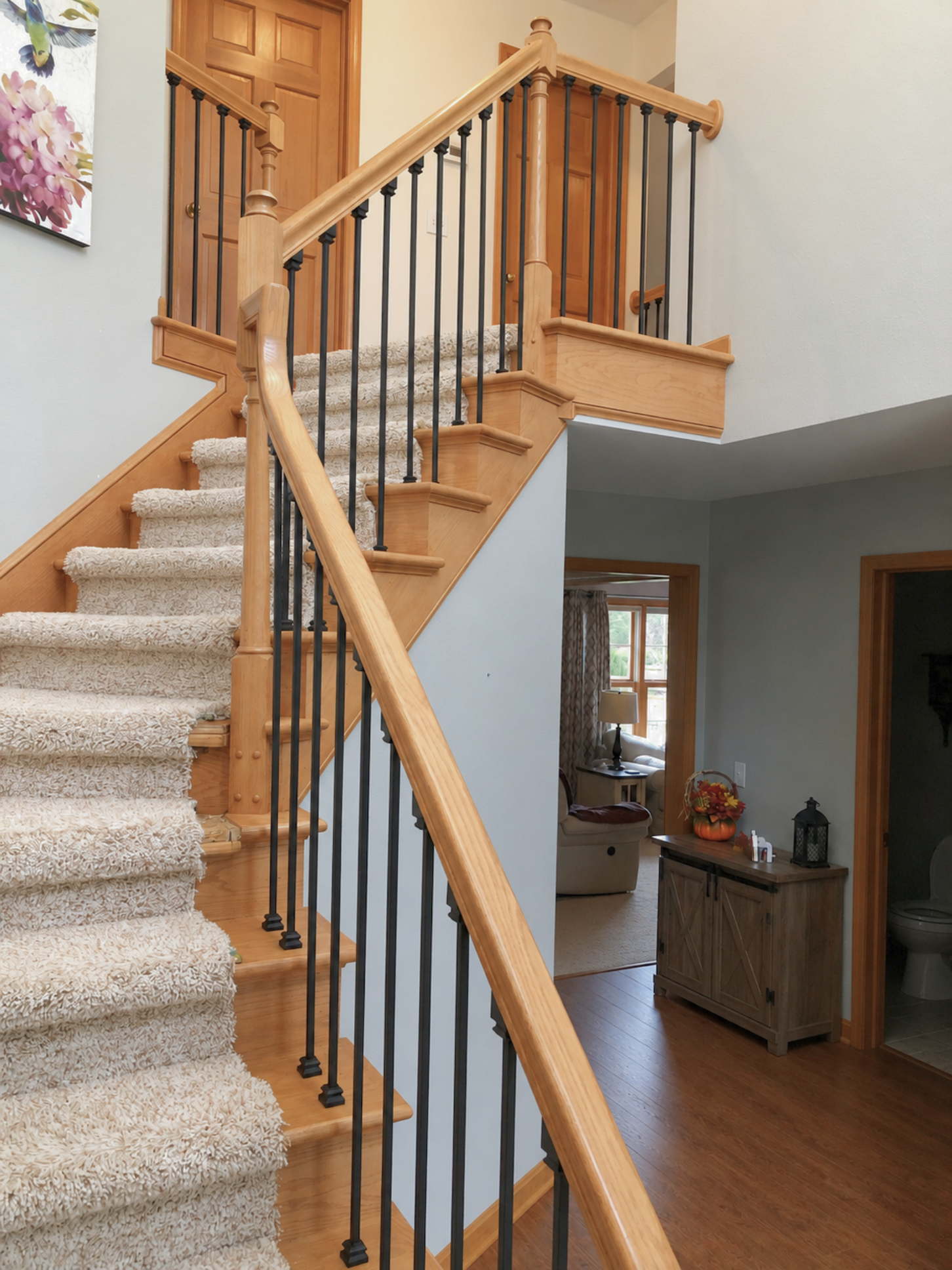 An indoor staircase with wooden steps covered by beige carpet, black metal balusters, and a wooden handrail, leading to a second floor with closed wooden doors. The area includes other rooms visible in the background, one with a window and a lamp, and another with a small cabinet decorated with pumpkins and flowers, and a lantern.