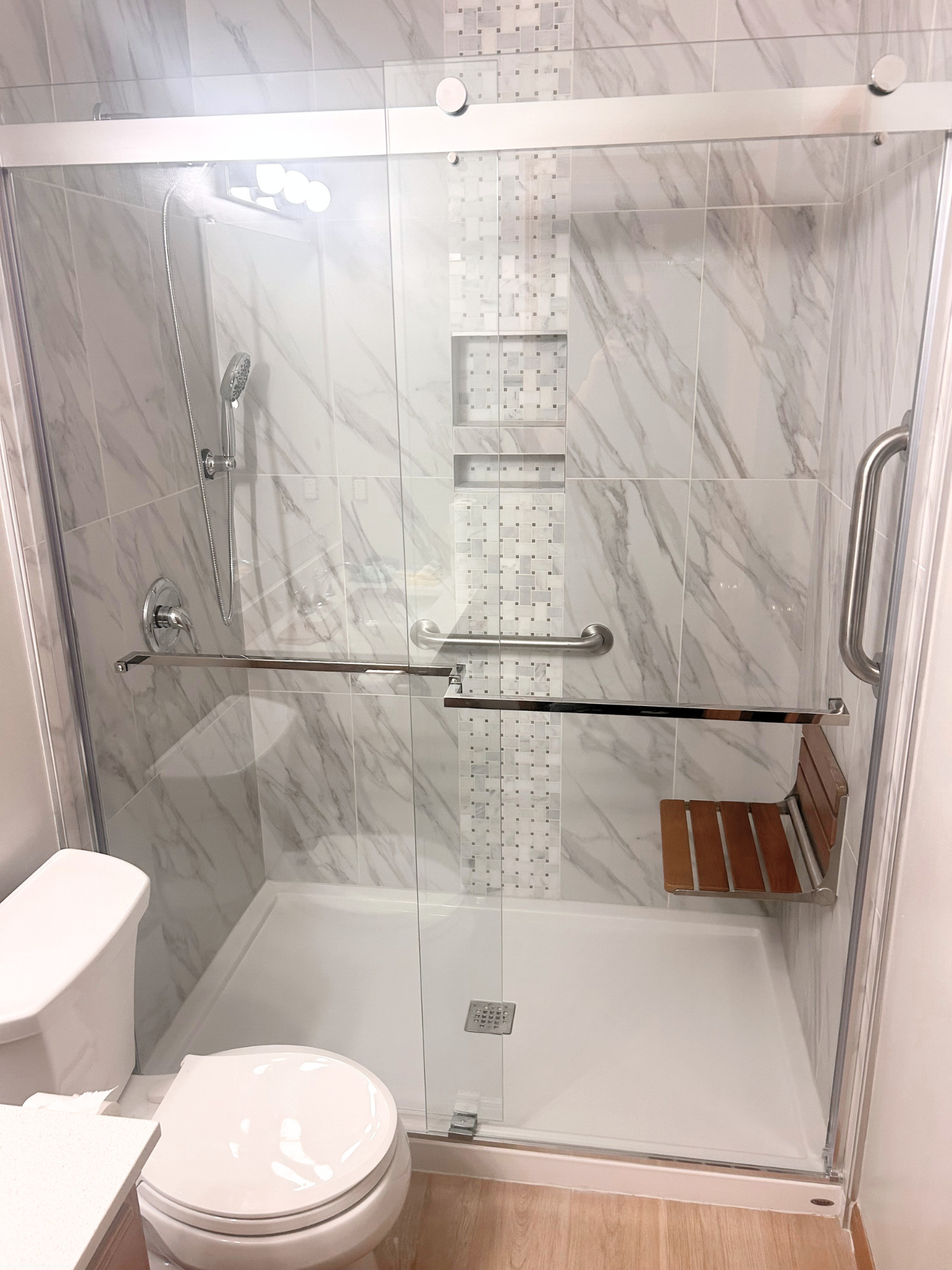 A bathroom shower area with glass doors, marble-look tiles, built-in shelves, a hand-held showerhead, metal grab bars, a white toilet, and a wooden bench.